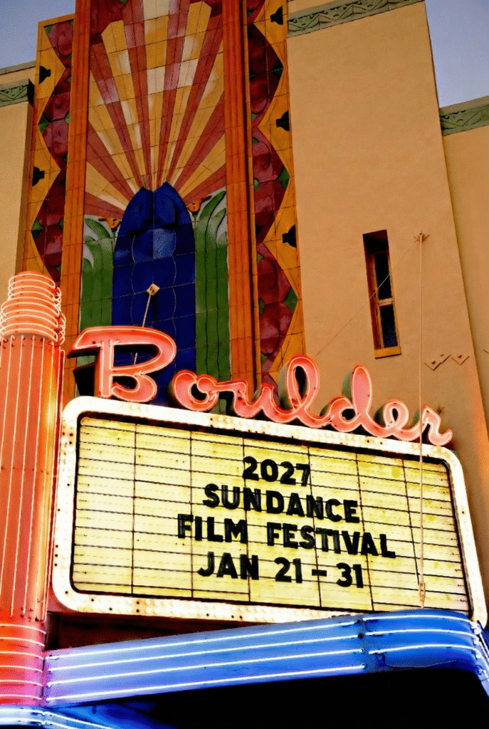 The theater's marquee displays information about the 2027 Sundance Film Festival, which runs from January 21 to 31. The theater's name, 'Boulder,' is illuminated in pink neon lights. The building features colorful, decorative artwork above the marquee.