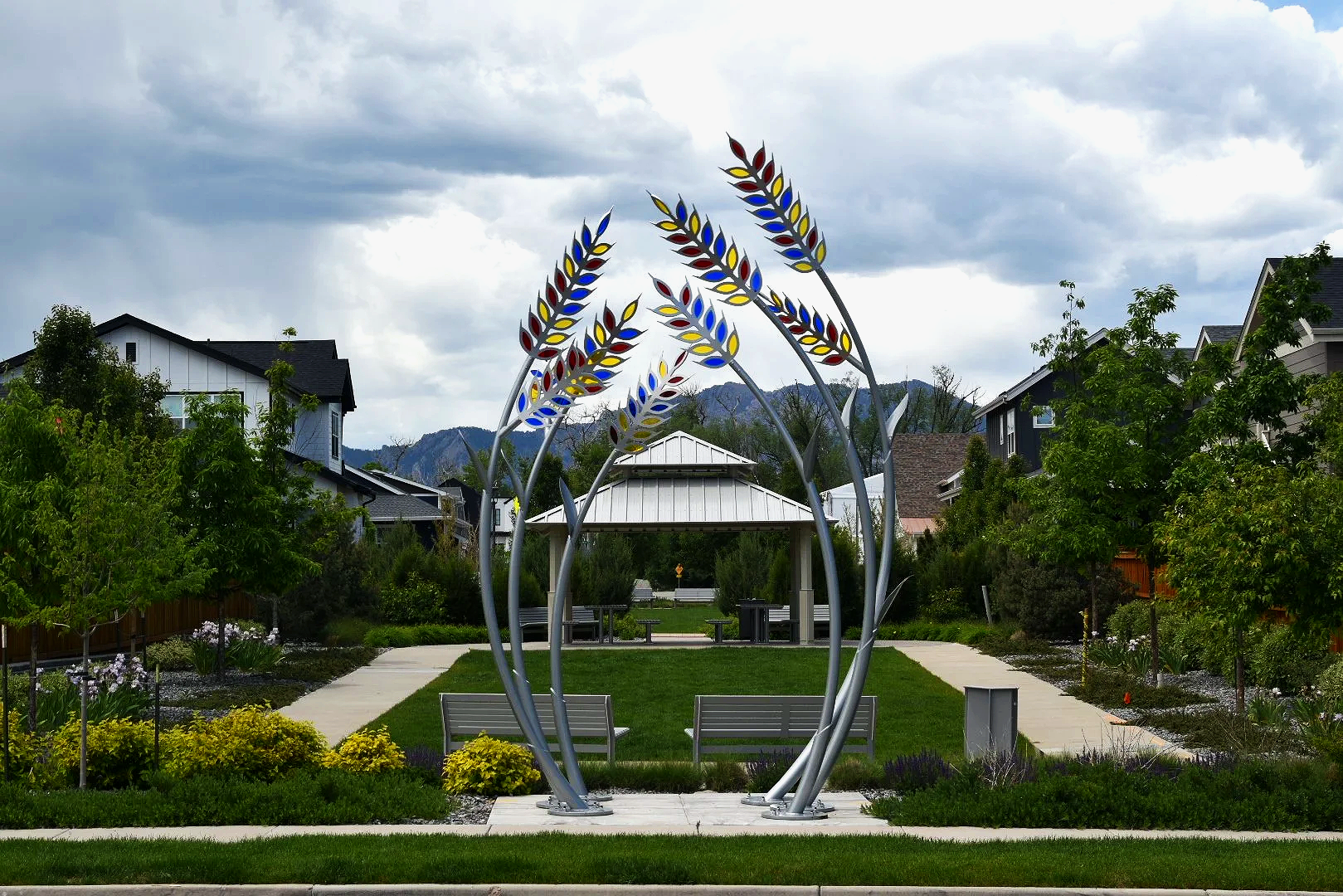 A park with a modern sculpture resembling stylized wheat stalks, benches, and a pavilion surrounded by greenery and houses, with mountains in the background under a cloudy sky.