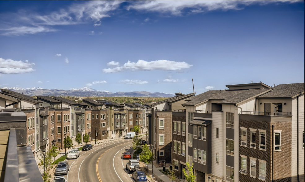 Residential street with modern apartment buildings, parked cars, and a mountainous landscape in the background under a partly cloudy sky.