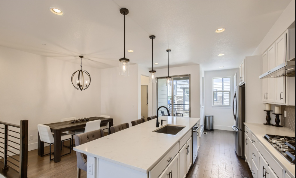 Modern kitchen with white cabinets, a large kitchen island with a sink, hanging pendant lights, and a dining area with a table and chairs.