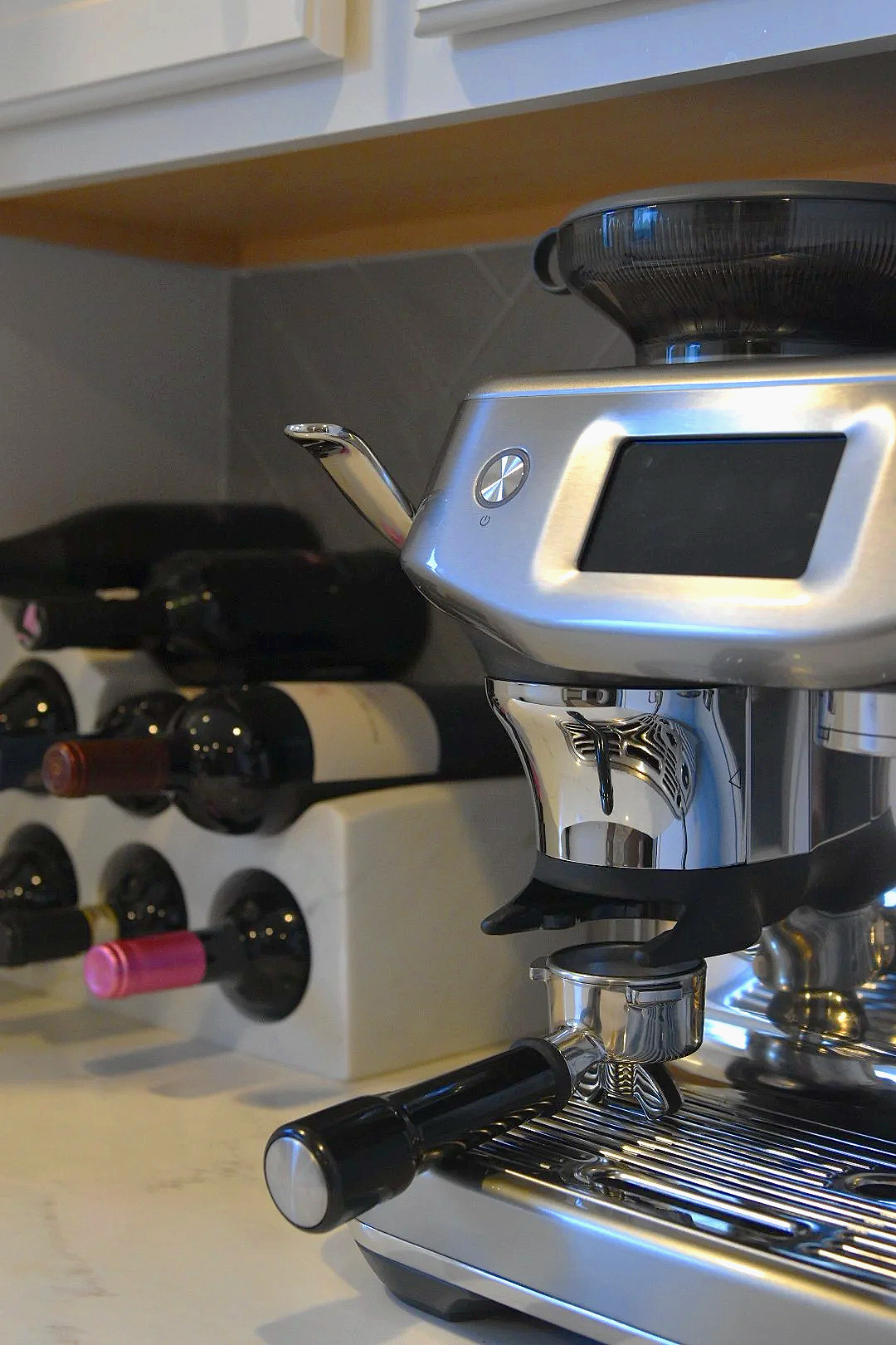 Close-up of a silver espresso machine on a kitchen counter with a black steam wand and a black handle. In the background, there is a white wine rack with several wine bottles stored horizontally.