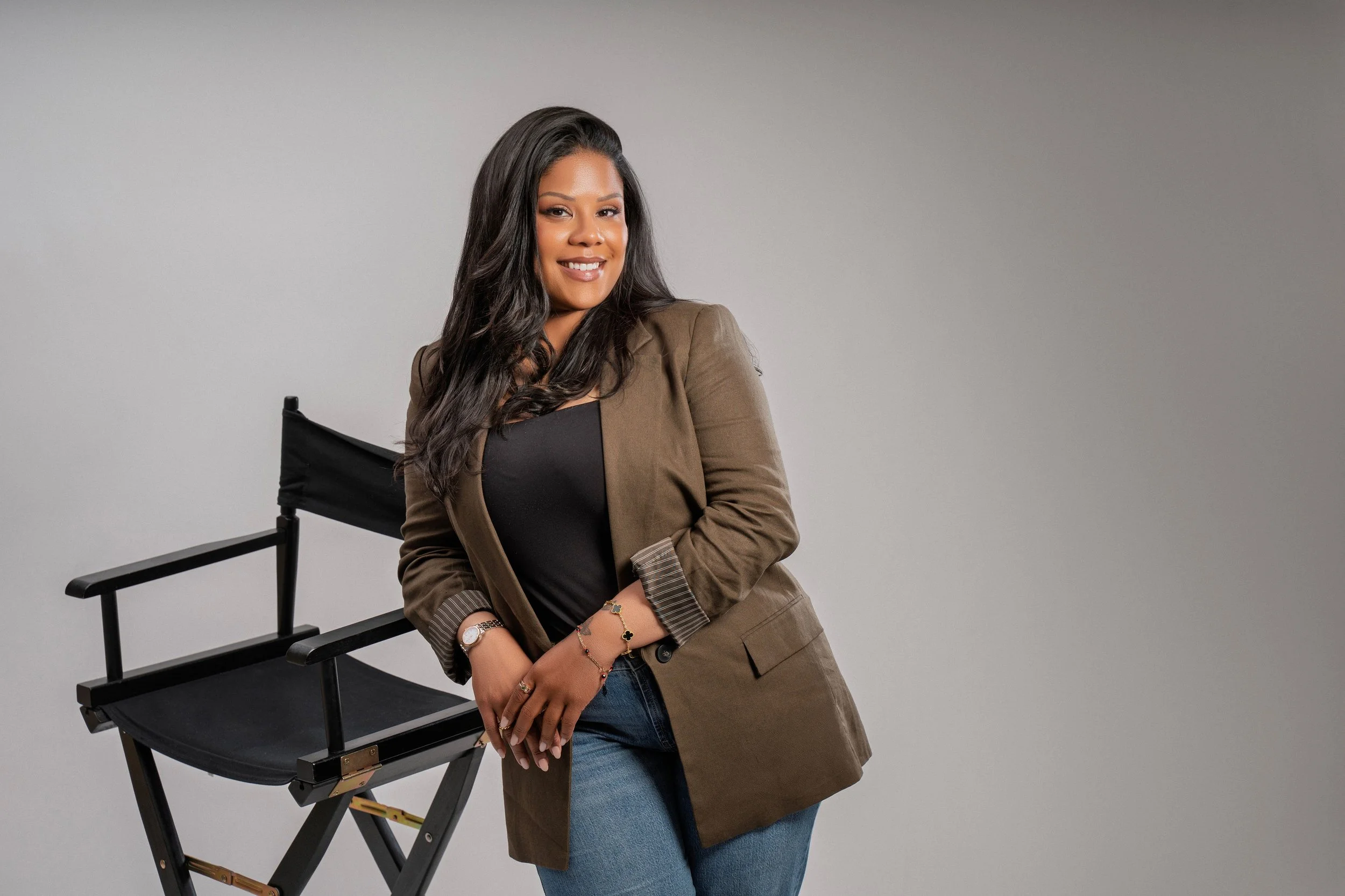 A woman with long dark hair, wearing a brown blazer over a black top and blue jeans, sitting beside a black director's chair, smiling at the camera against a plain gray background.