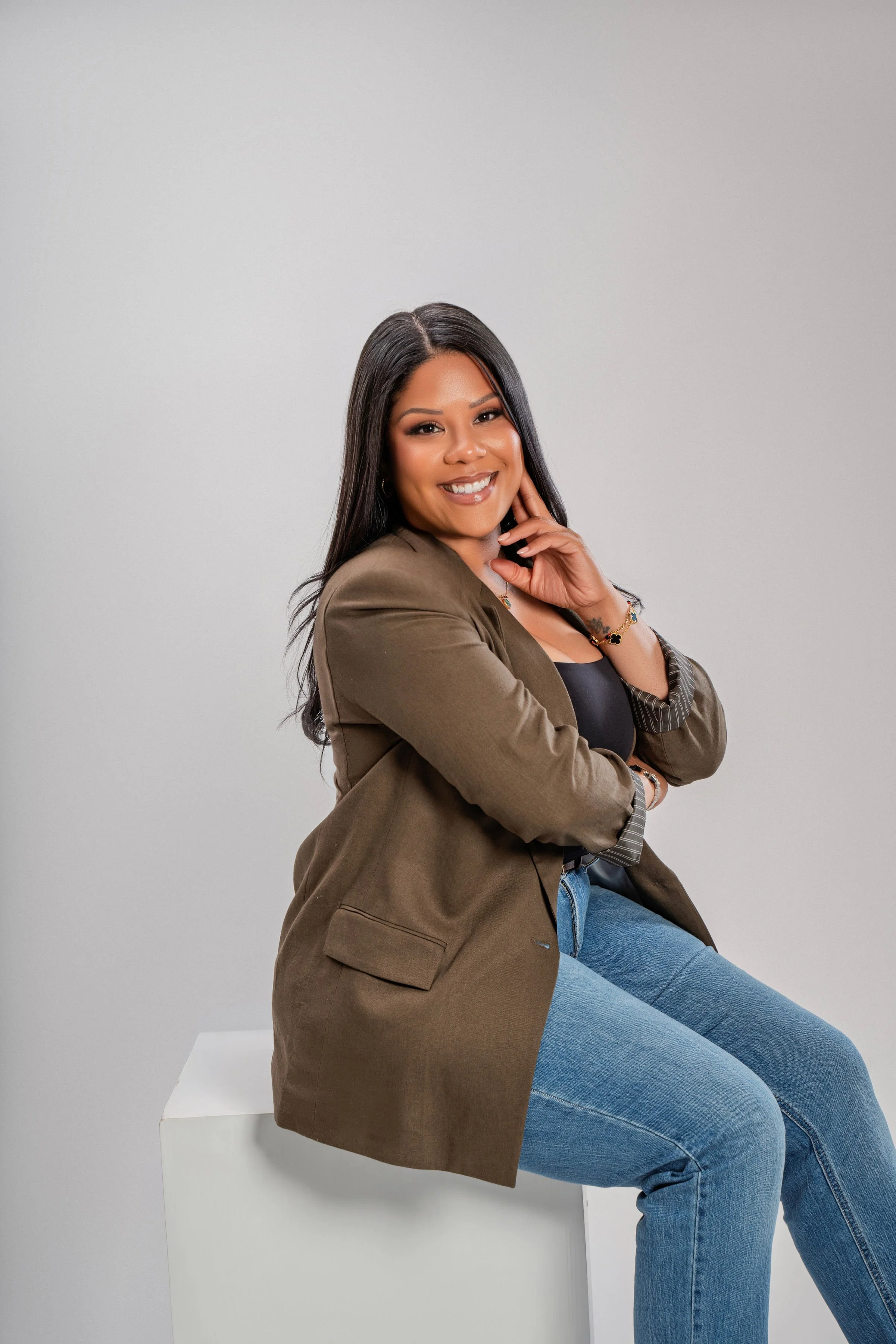 A woman with long dark hair, wearing a brown blazer, black top, and blue jeans, sitting on a white block against a light gray background. She is smiling and resting her chin on her hand.