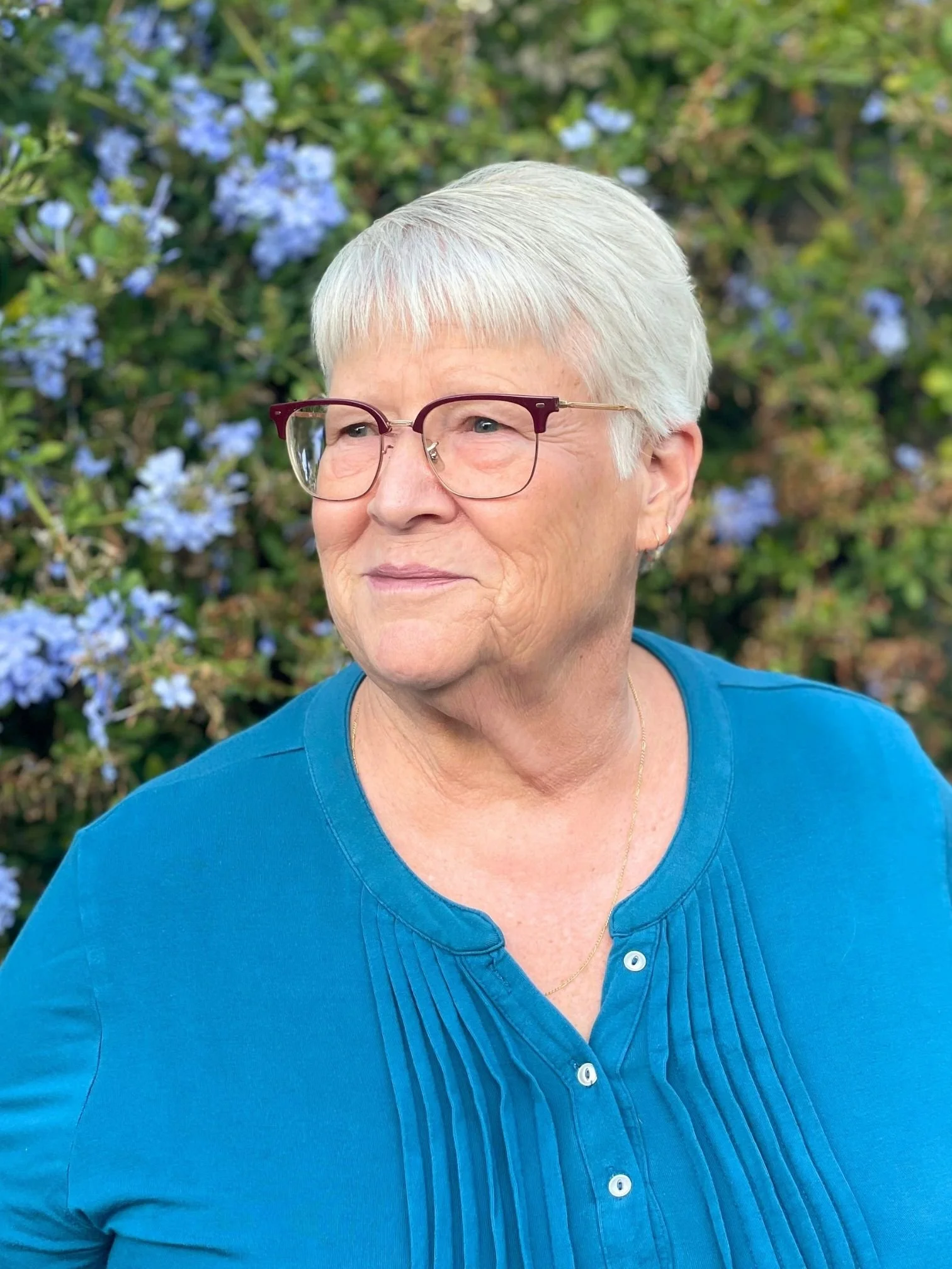 Close-up of Ms. Louise Thompson from LL Science Tutoring wearing a blue top, standing outdoors in front of green foliage with light blue flowers.