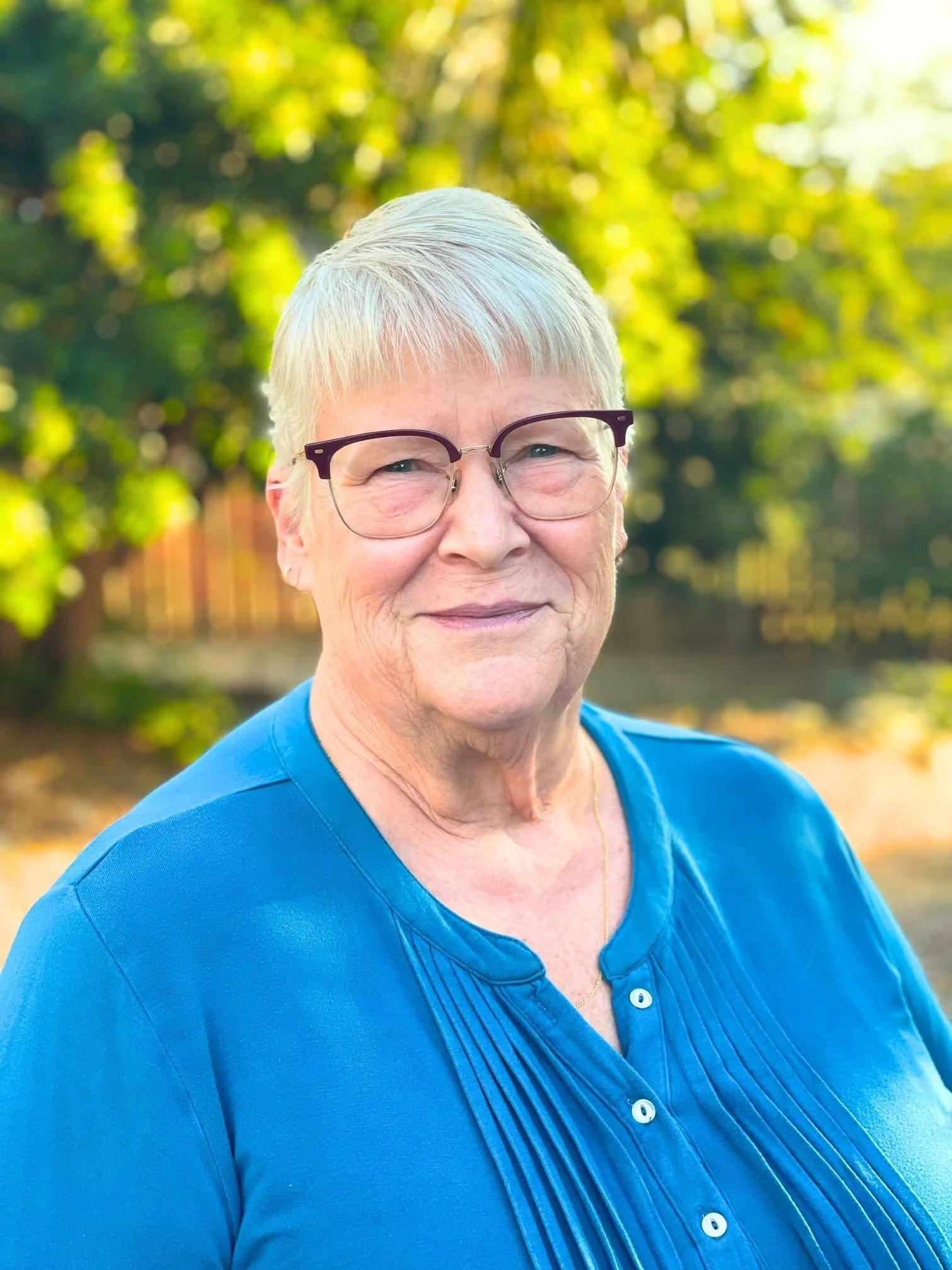 Close-up of Ms. Louise Thompson from LL Science Tutoring smiling, outdoors with green trees in the background, wearing a blue shirt.