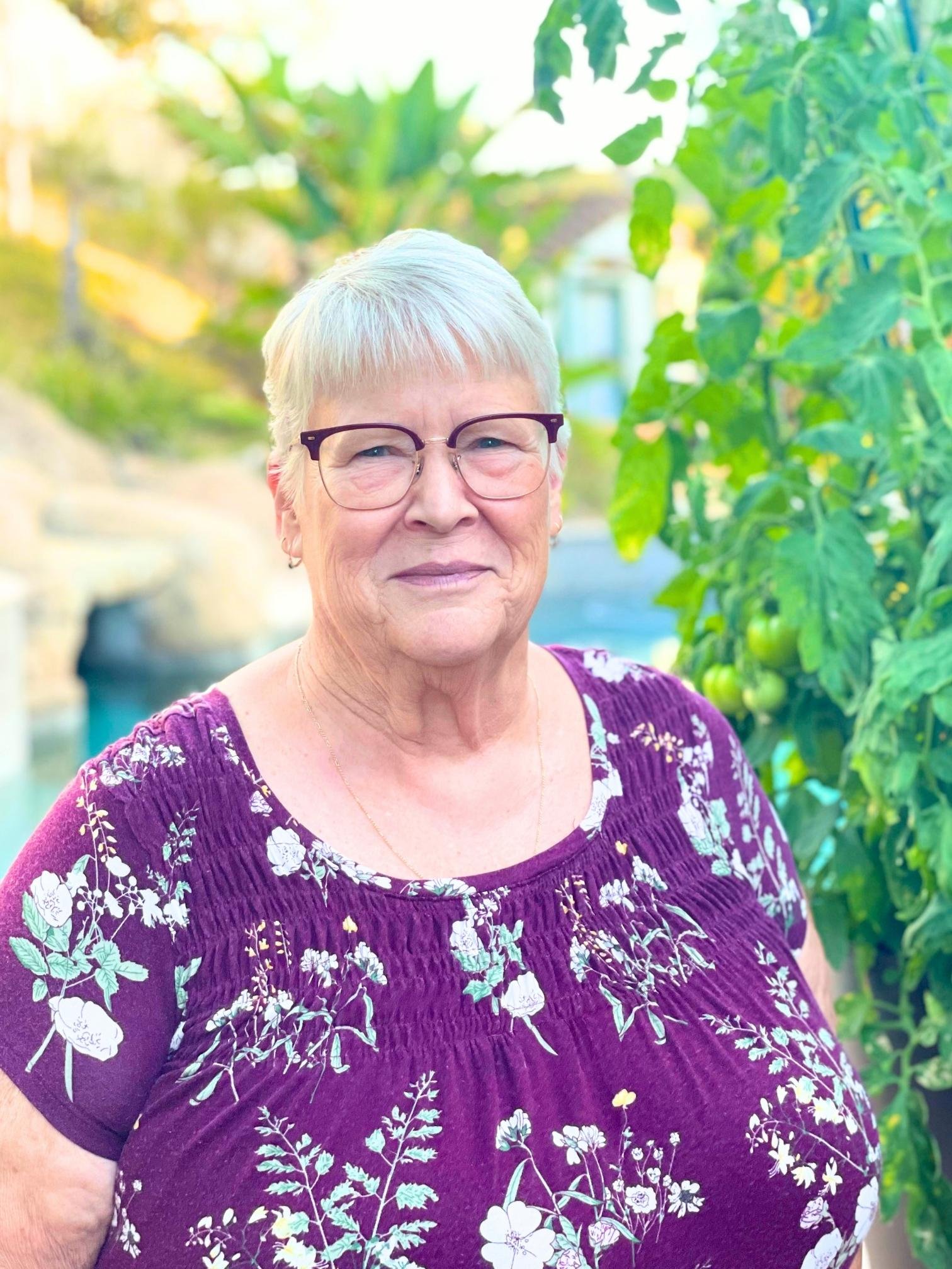 Close-up of Ms. Louise Thompson from LL Science Tutoring wearing glasses, and a purple floral dress, standing outdoors next to green leafy plants.