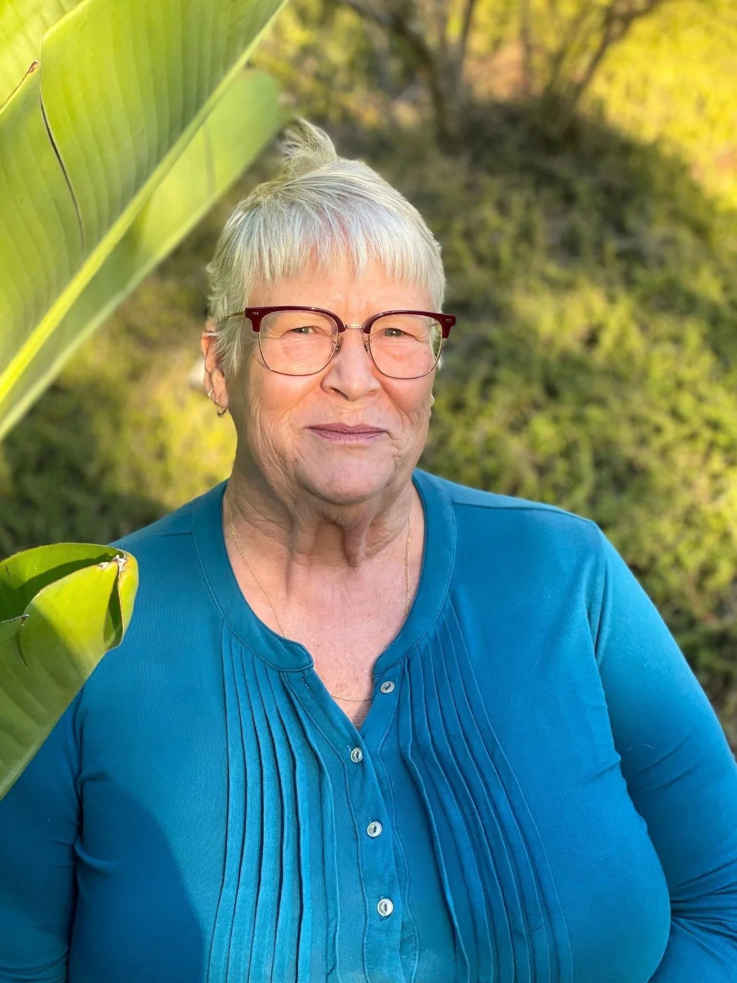 Close-up of Ms. Louise Thompson from LL Science Tutoring wearing glasses and a blue shirt, standing outdoors with blurred green foliage in the background.
