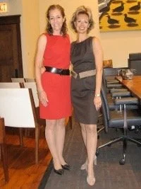Two women in dresses standing in a room with a dining table and chairs, smiling at the camera.