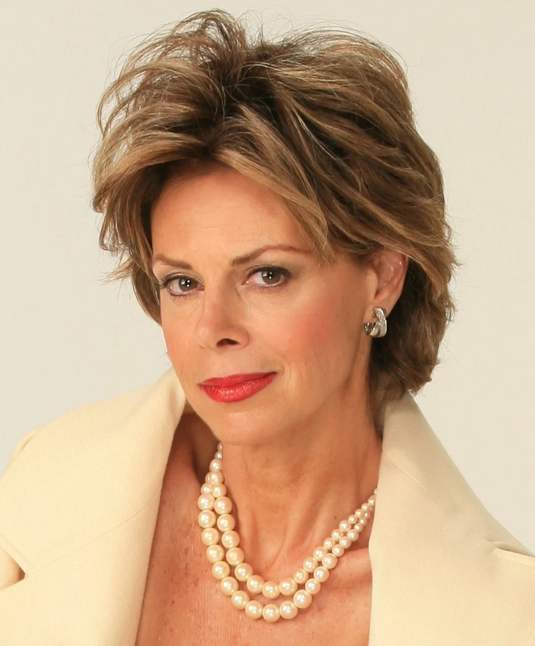 A woman with short, styled brown hair wearing pearl earrings, a pearl necklace, and a cream-colored blazer, posing against a plain light background.
