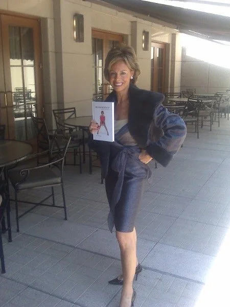 Woman standing outdoors on a patio, smiling, holding a magazine with a person in red on the cover, dressed in a dark blazer and skirt, with tables and chairs in the background.