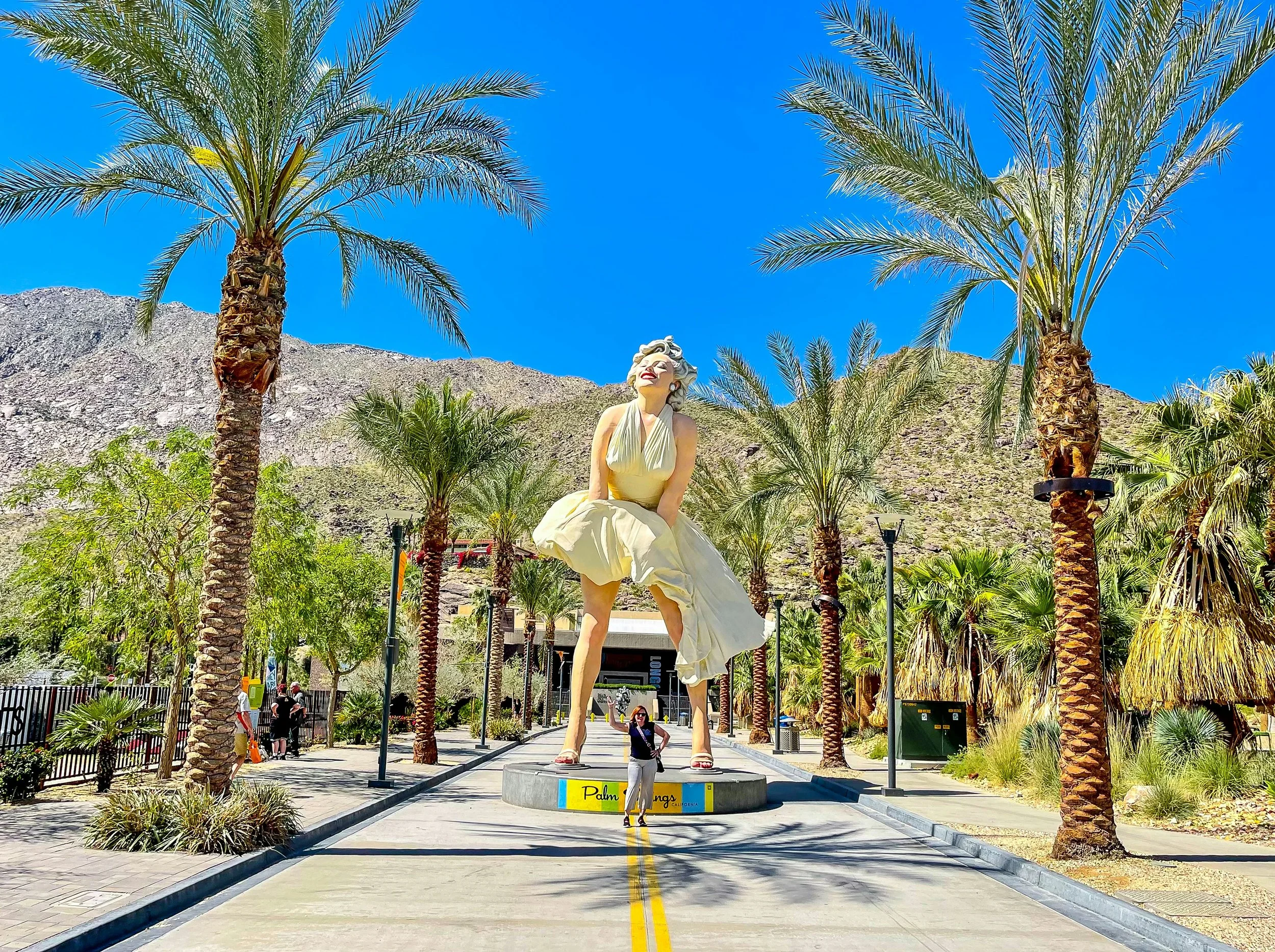 A large Marilyn Monroe statue dressed in a white dress with a gust of wind, located in Palm Springs, California, surrounded by palm trees and people.