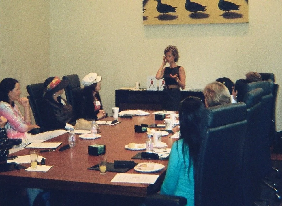 A woman standing at the head of a conference table giving a presentation to seven seated women and men in a professional setting. The room has beige walls with a painting of black ducks on a yellow background.