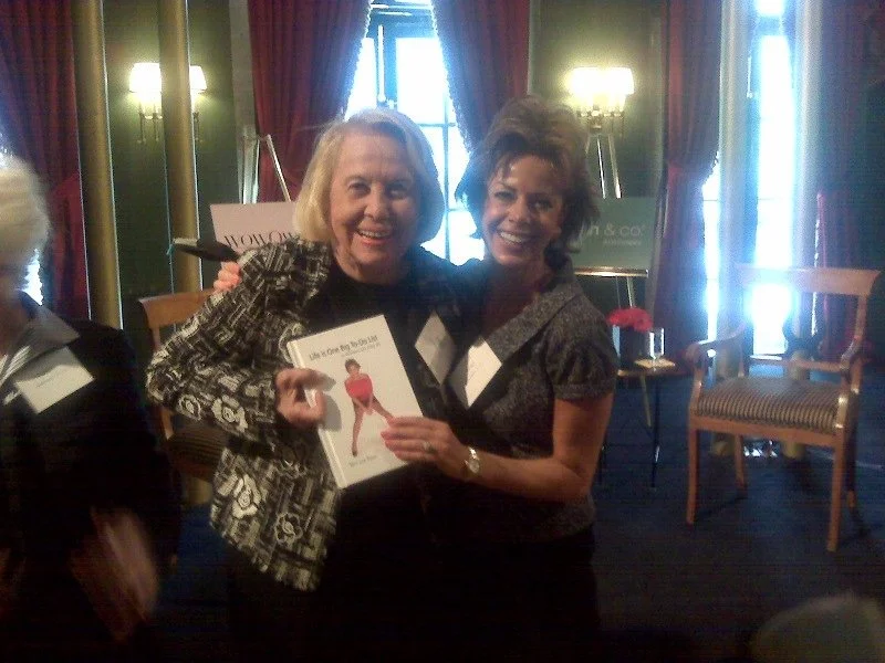 Two women smiling and holding a book in a decorated room with curtains, chairs, and lamps.