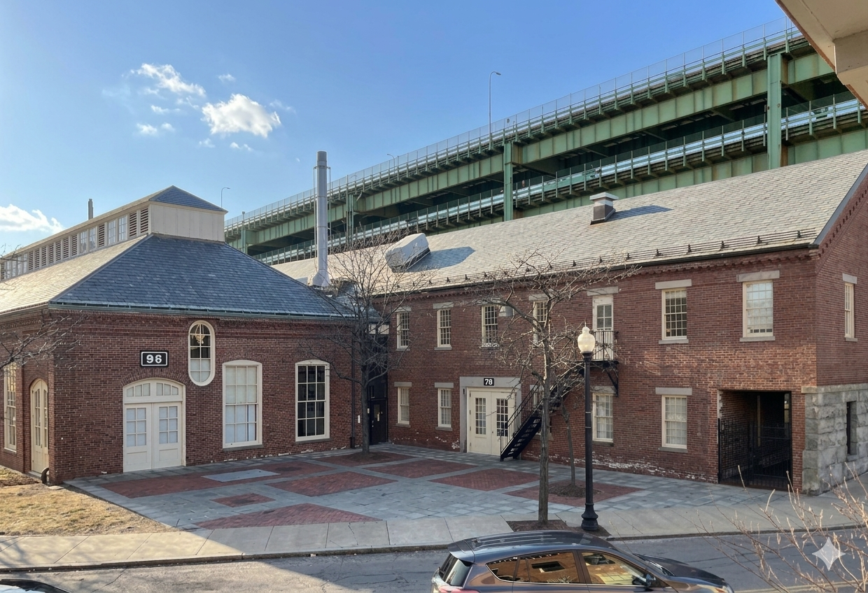 Two brick buildings with arched and rectangular windows and doors, a paved courtyard, leafless trees, a streetlamp, and a parked car, with an elevated highway in the background under a partly cloudy sky.