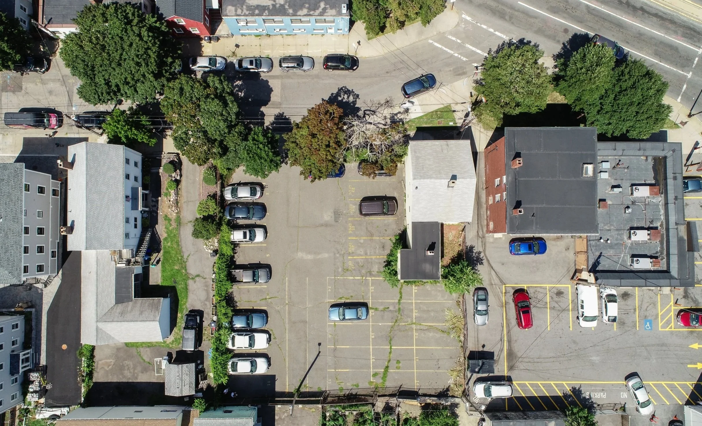 An aerial view of a city street with parked cars, trees, and buildings. There is a parking lot with numbered spaces and a few cars on the street.