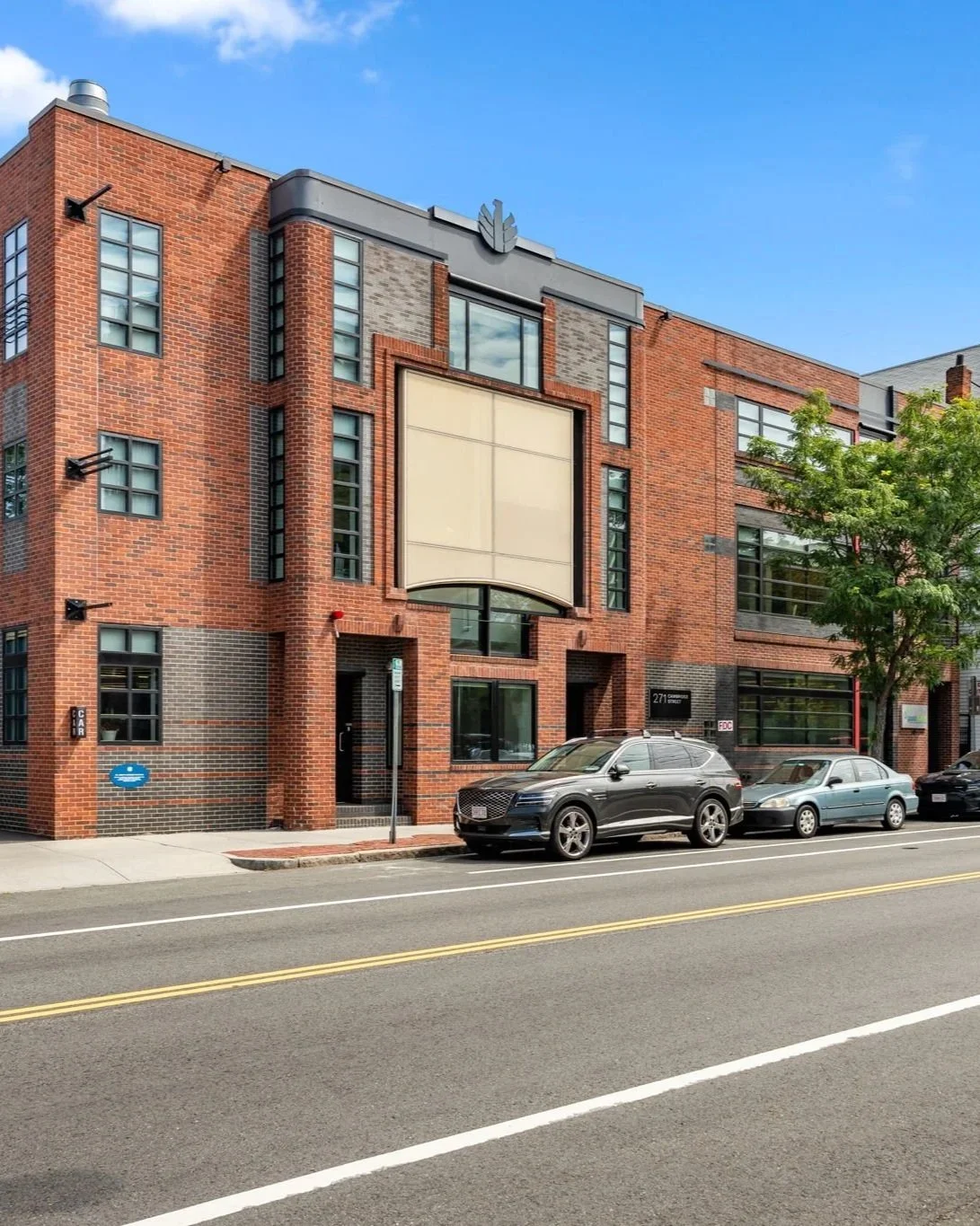 A red brick building with large windows and a blank billboard on the front, parked cars along the street, and a tree in front with a clear blue sky overhead.