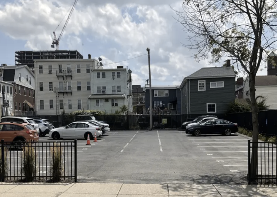 A parking lot with several parked cars, traffic cones, and a fenced entrance, with residential buildings and a construction crane in the background under a partly cloudy sky.