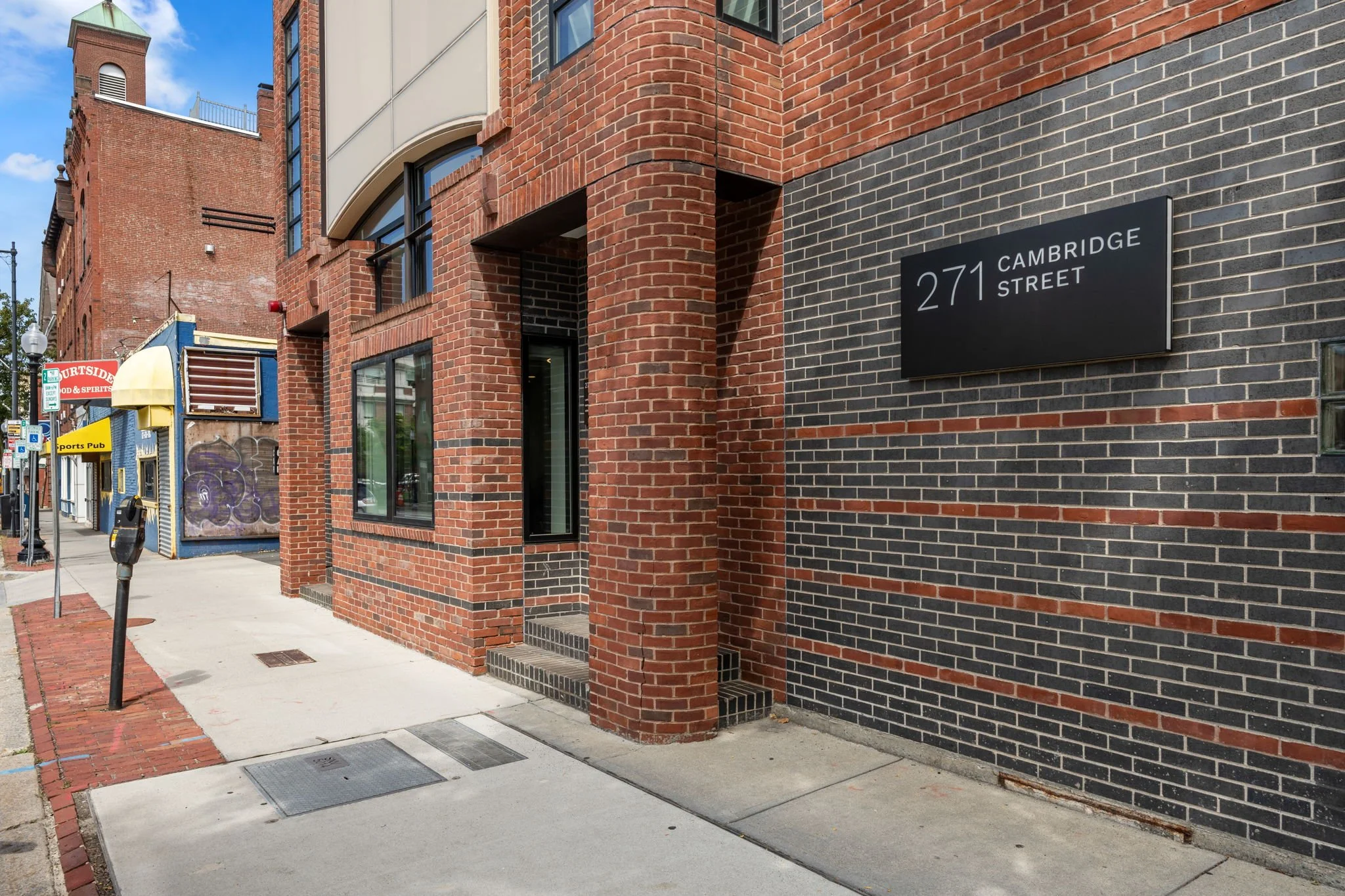 A city sidewalk next to a brick building with a black and red brick pattern. A street sign indicates Cambridge Street and there are storefronts and a parking meter in the background. The sky is partly cloudy.
