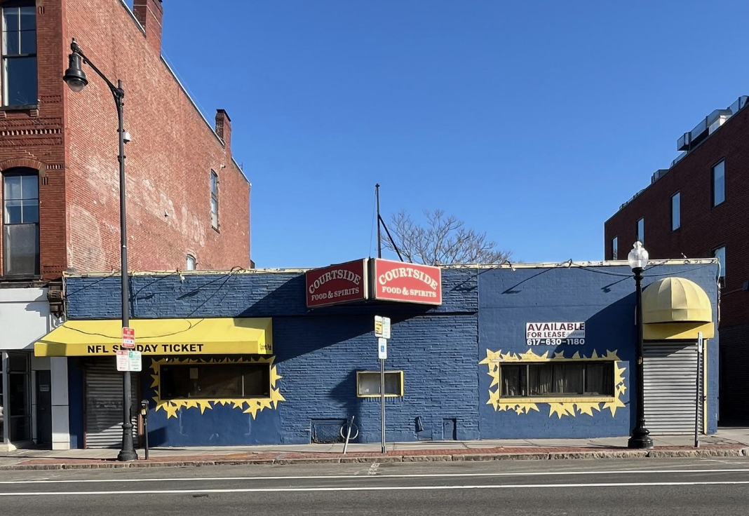 A small blue storefront with yellow awnings and yellow star border decals, signage for Courtside Food & Spirits, a NFL Sunday Ticket advertisement, and an 'Available for Lease' sign with a phone number.