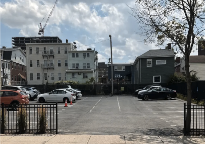 Empty outdoor parking lot with a few parked cars, surrounded by a black fence, with multi-story buildings and a construction crane in the background under a partly cloudy sky.
18 Winter St. 