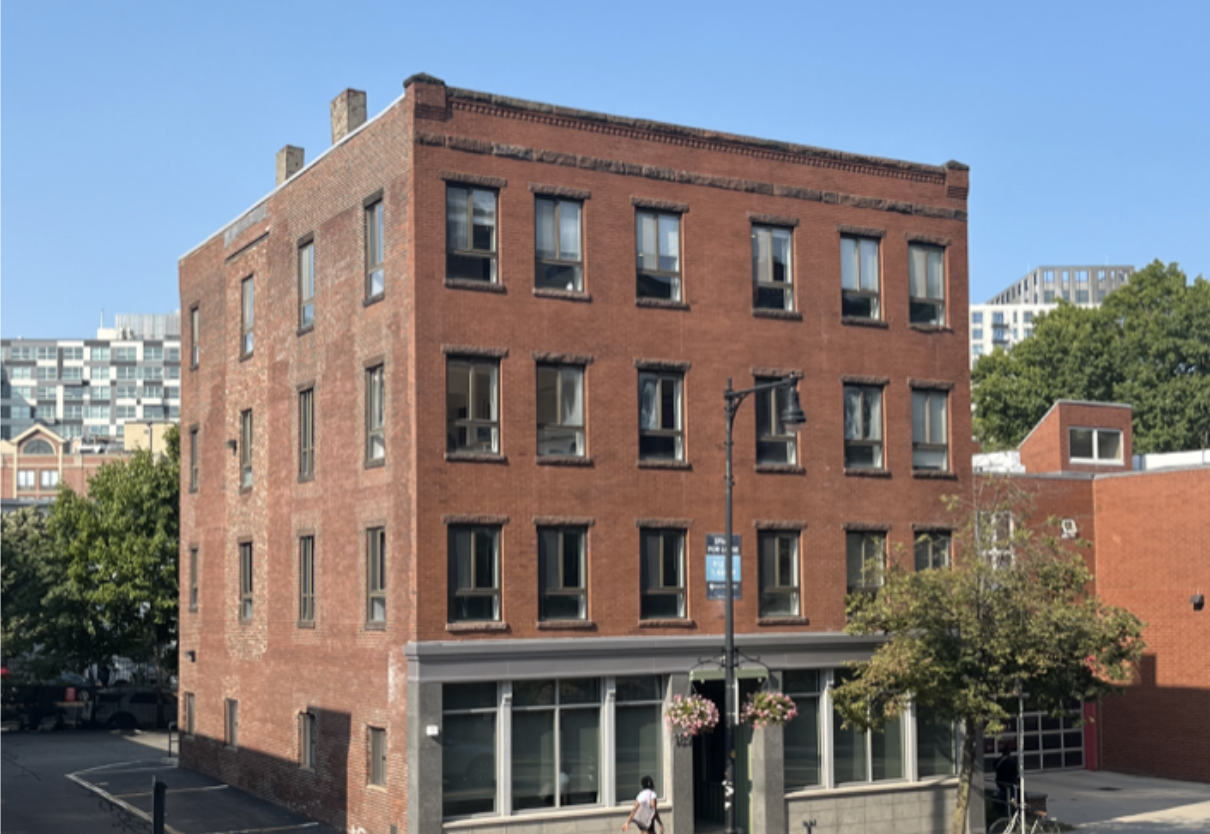 A multi-story red brick building with large windows and a store on the ground floor, situated on a street corner with a lamp post, street signs, hanging flower baskets, trees, and a person walking by on a sunny day.