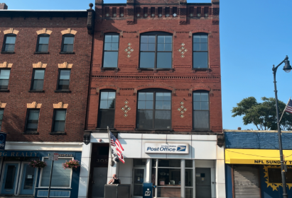 Three-story red brick building with large windows, located on a city street with a Post Office on the ground floor, American flags, and a streetlamp. Adjacent buildings are also visible.