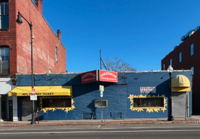 Downtown storefront with blue and yellow exterior, including for sale and NFL ticket signs, and an American flag on a lamppost.