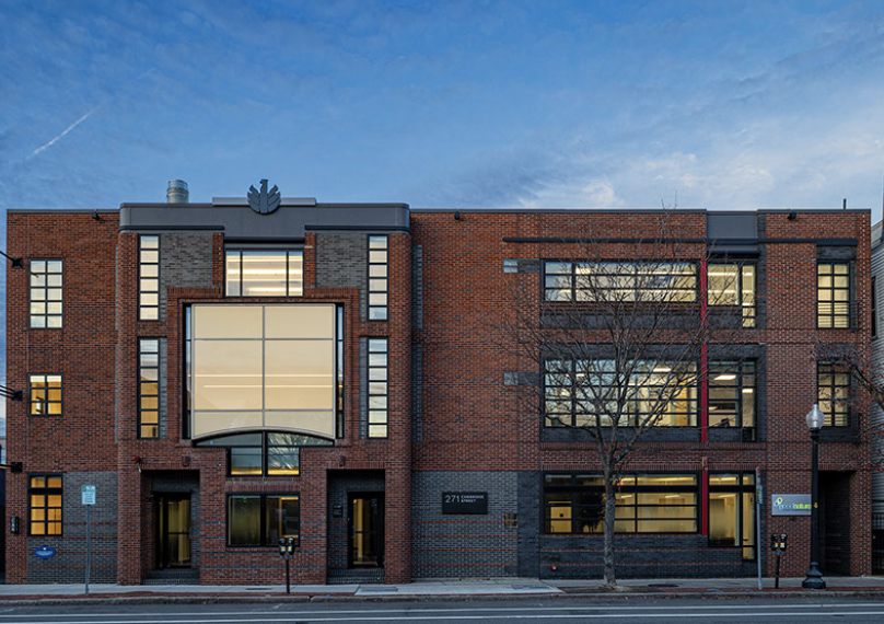 Exterior view of a modern brick office building at dusk, with illuminated windows and a prominent glass entrance, street and tree in front. 271 Cambridge St.