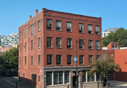 A four-story red brick building with large windows on each floor, a small awning at the entrance, and hanging flower baskets, situated on a city street corner with trees and other buildings nearby.