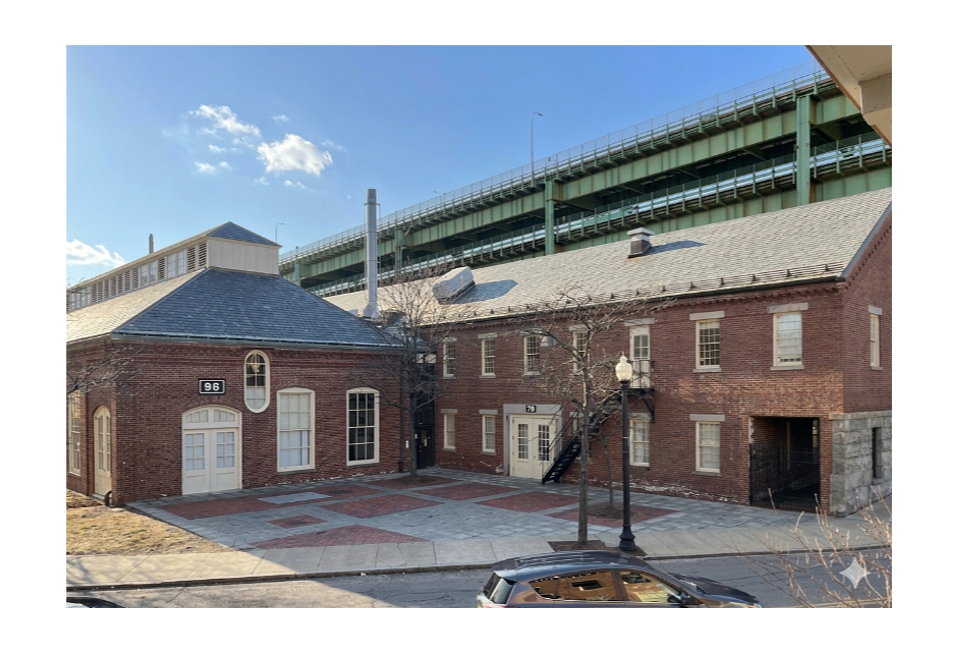 Urban scene showing a brick building with white trim and a black staircase, a leafless tree, a sidewalk, a street with a car, and an elevated highway above the building against a blue sky with a few clouds.