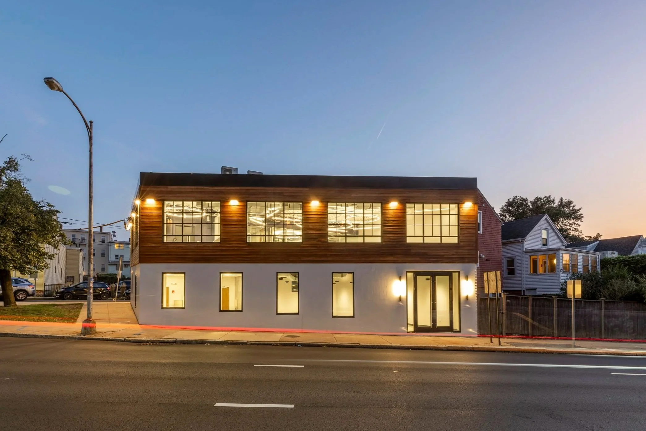 Modern two-story building with wood paneling and large black-framed windows, three small planted trees on the sidewalk, a one-way street sign, and a street address sign reading '15 Winter Street' on a cloudy day.
