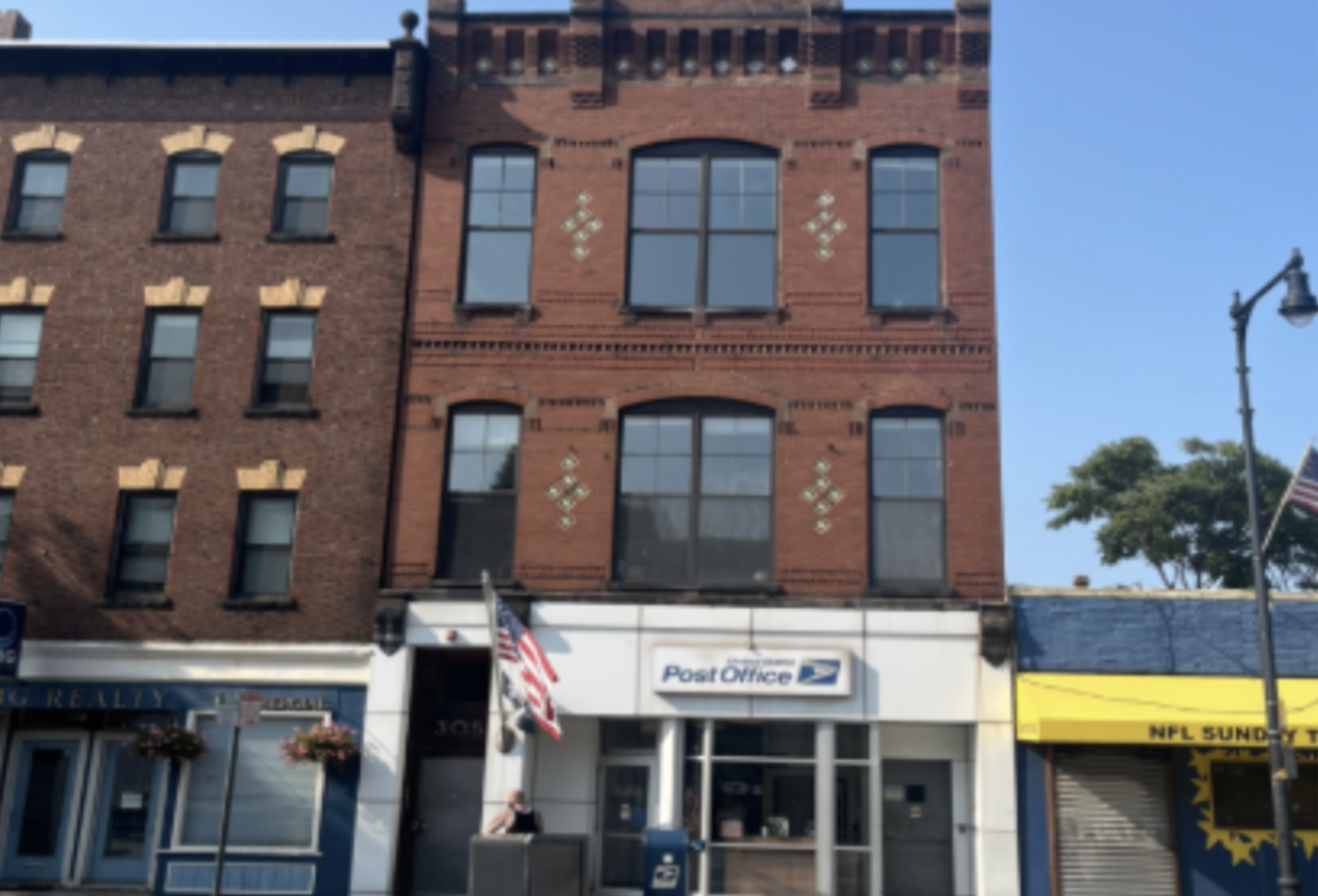 A city street scene showing three buildings with retail storefronts on the ground floor. The middle building, made of red brick, houses a post office, and the building on the right has a yellow and blue sign. There are American flags hanging outside the stores, and a street lamp on the right side of the image. The sky is clear and blue.