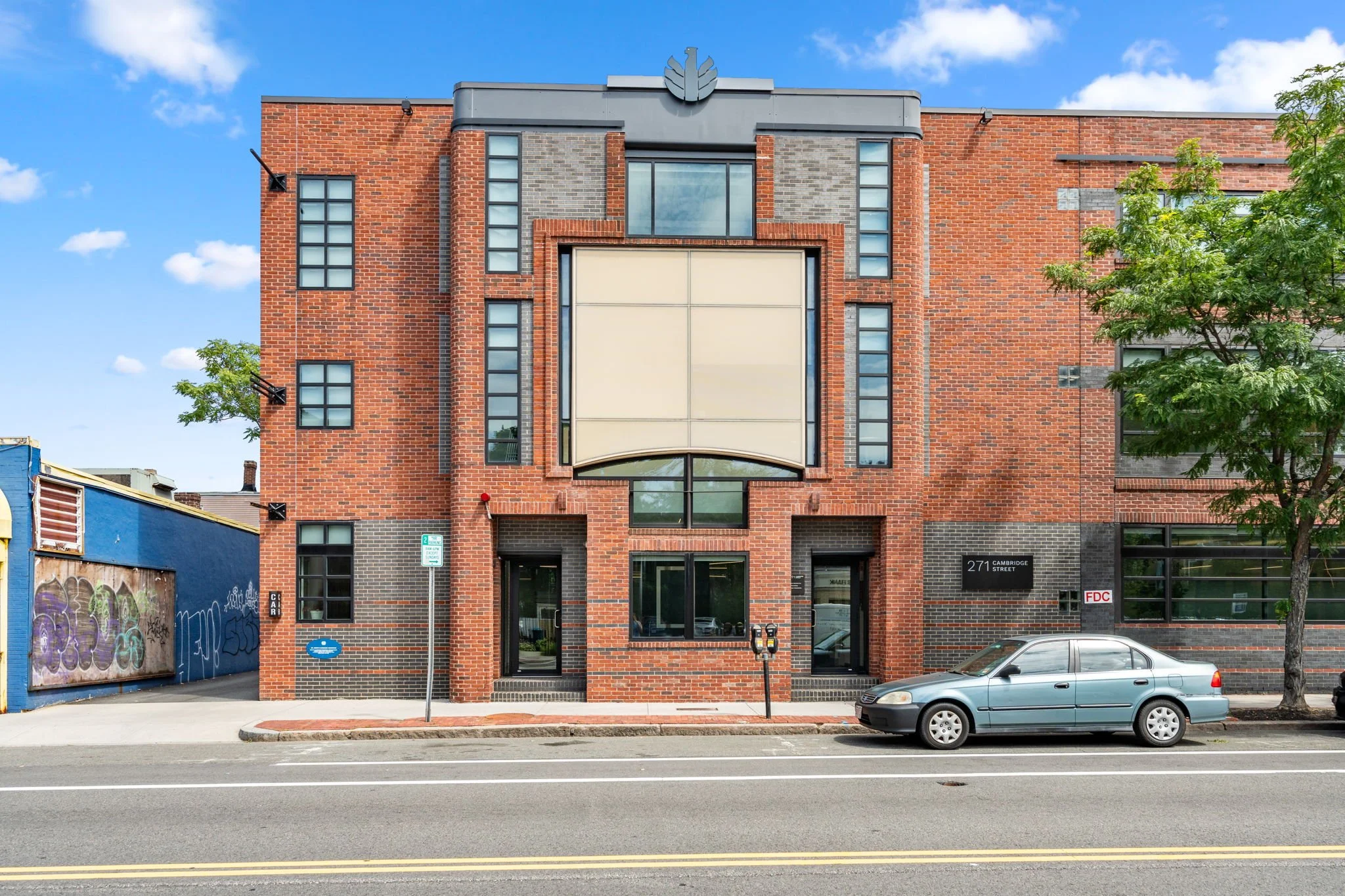 A red brick building with multiple rectangular windows, some with glass blocks, and a large beige sign or billboard in the center. There are trees on the right and what appears to be graffiti on a blue wall on the left. A silver car is parked in front of the building, and a sign indicating parking regulations is visible. The sky is blue with some clouds.