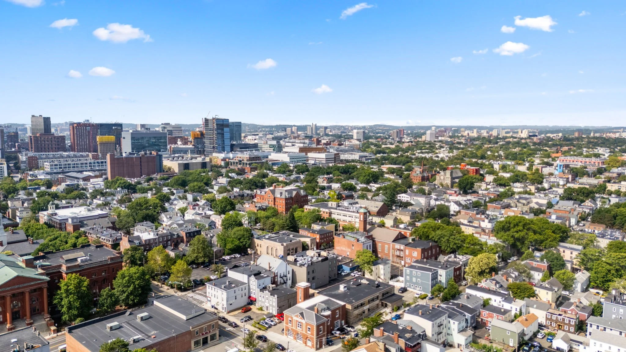 A city skyline with a mix of tall modern buildings and smaller residential houses, under a blue sky with scattered clouds.
