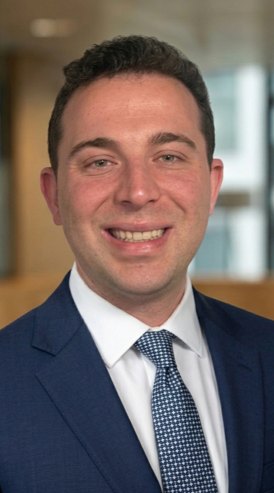 A young man with short dark hair and light skin, smiling in a professional setting, dressed in a navy suit, white shirt, and patterned tie. William Wiener. Wil.