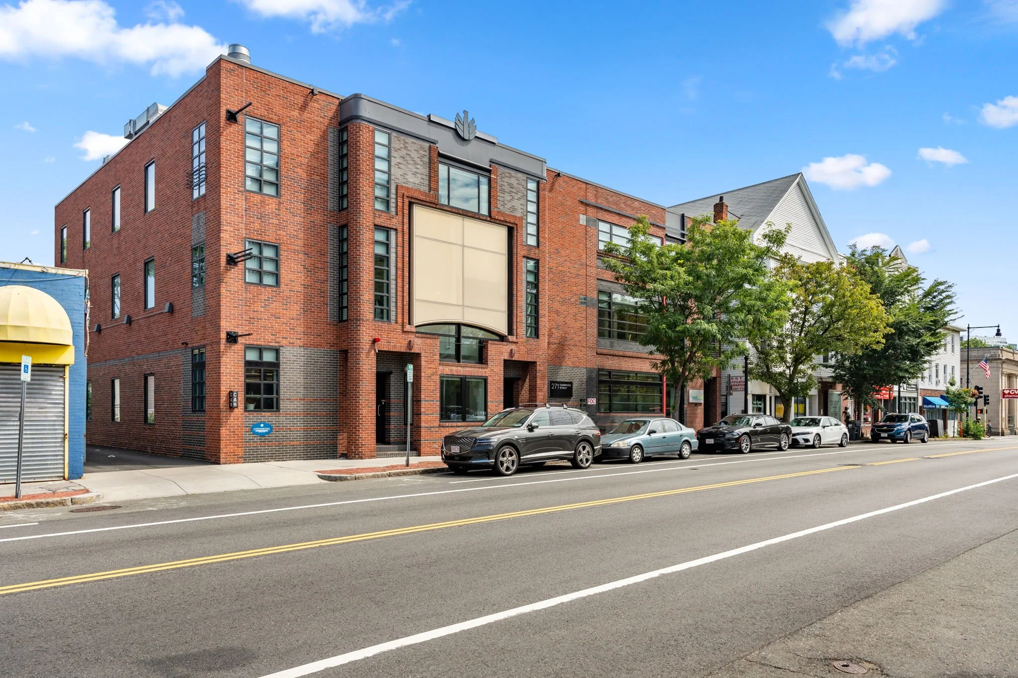 A street view of a multi-story red brick building with large windows, parked cars, a tree, and storefronts on both ends, under a partly cloudy blue sky.