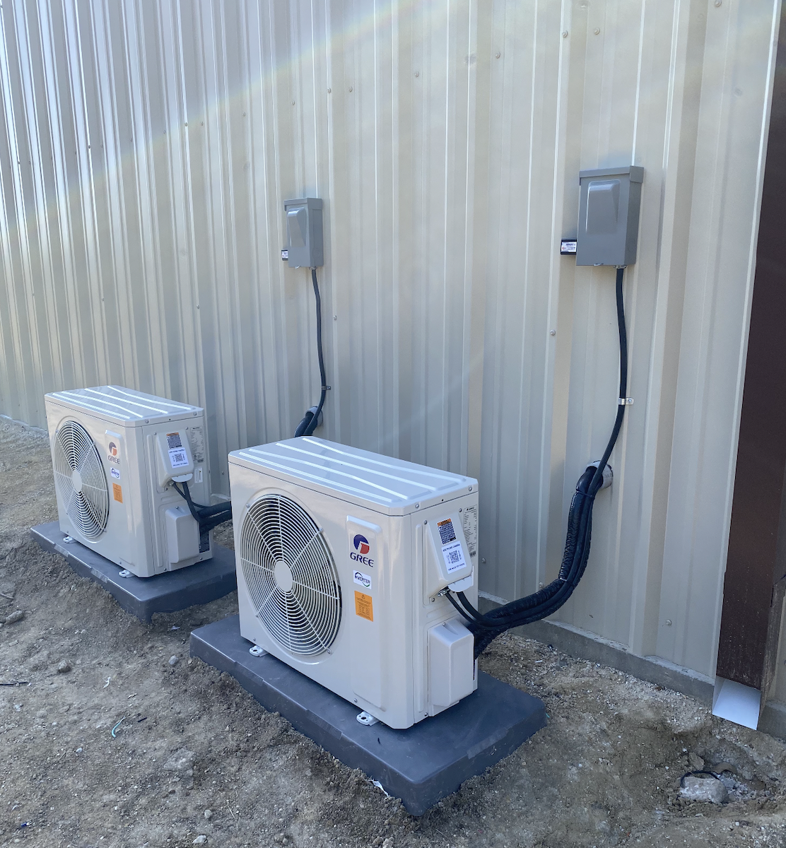 Two outdoor air conditioning units installed next to a metal wall with electrical boxes above.