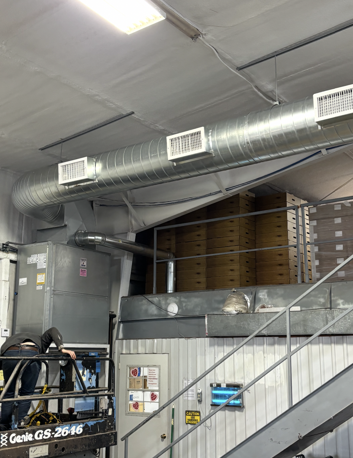 Industrial interior with large metal ventilation duct running along the ceiling, a worker on a lift, and stacked wooden boxes.