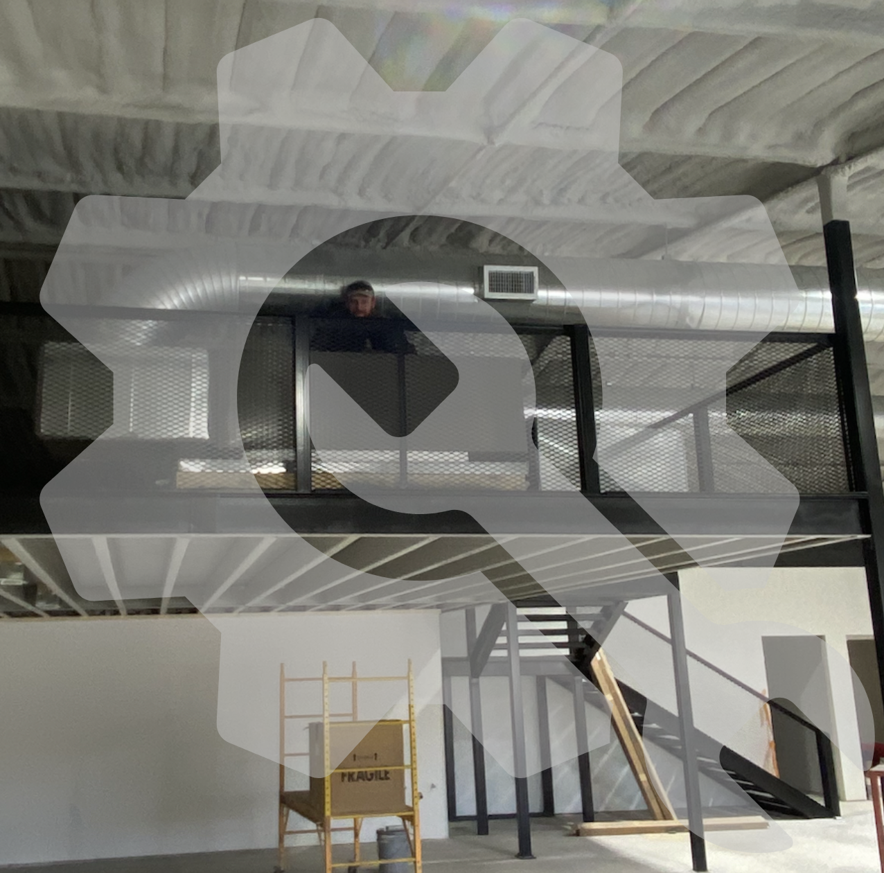 A construction site with a two-story metal structure. The upper level has a black railing and a person is standing behind it, peering over. There is a stairway leading to the upper level and construction materials including a yellow mobile scaffold labeled "FRAGILE" and a wooden plank are visible in the lower area.