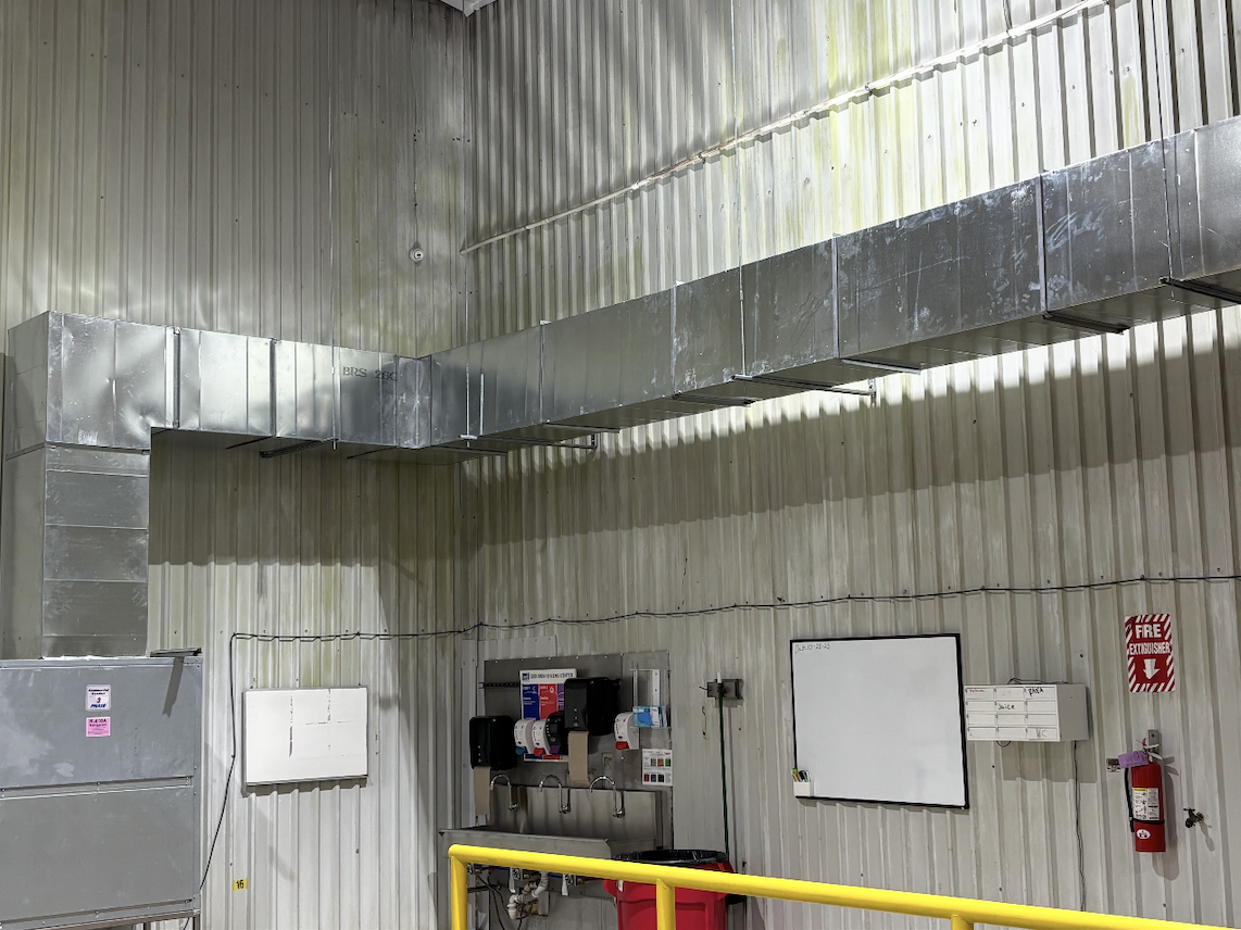 Interior of an industrial or commercial building with metal walls, large metal air ductwork running along the ceiling, a whiteboard, a fire extinguisher, and various safety and utility equipment.