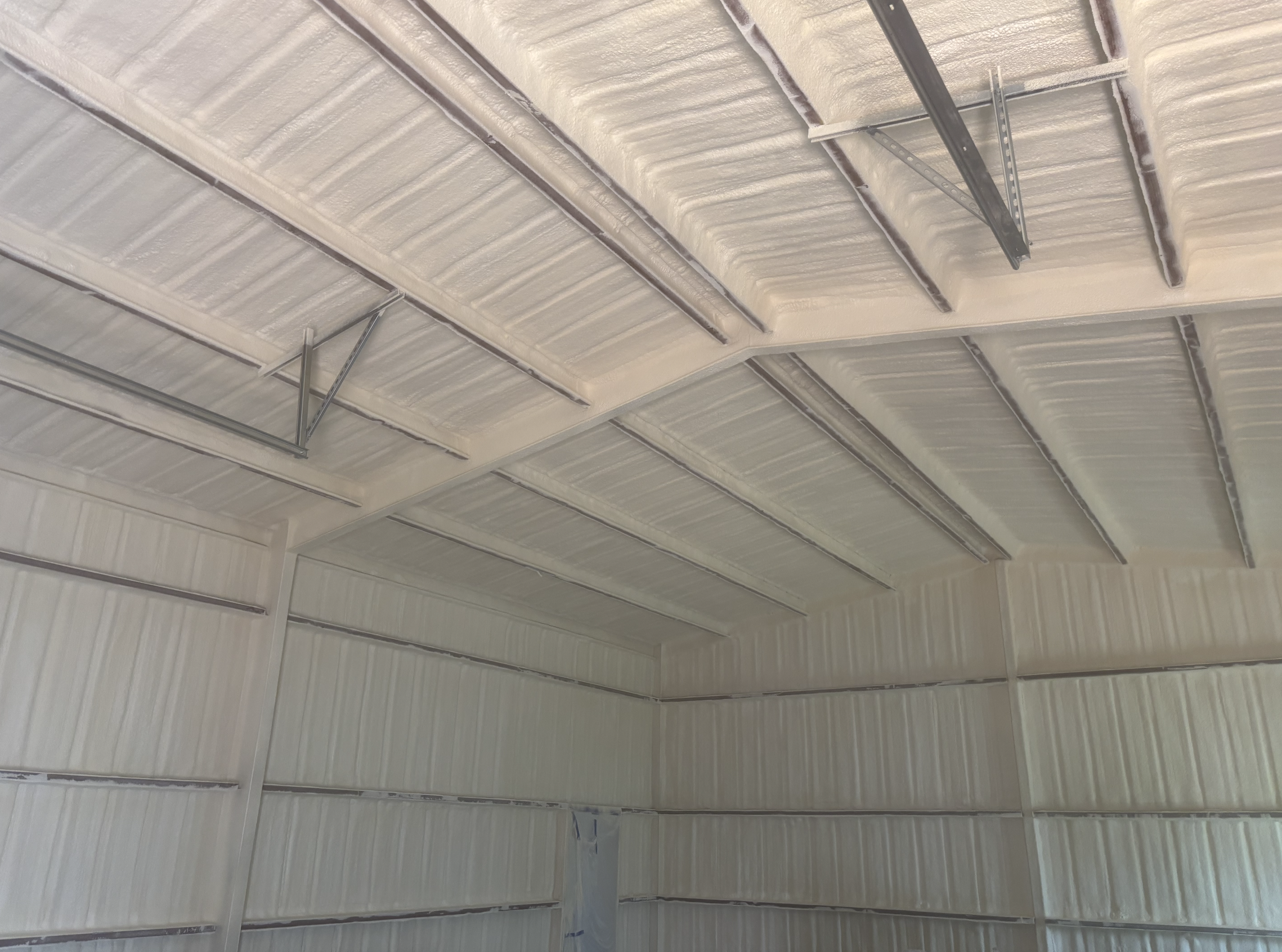 The image shows the ceiling of a building with insulation spray foam applied between exposed metal beams and framing.