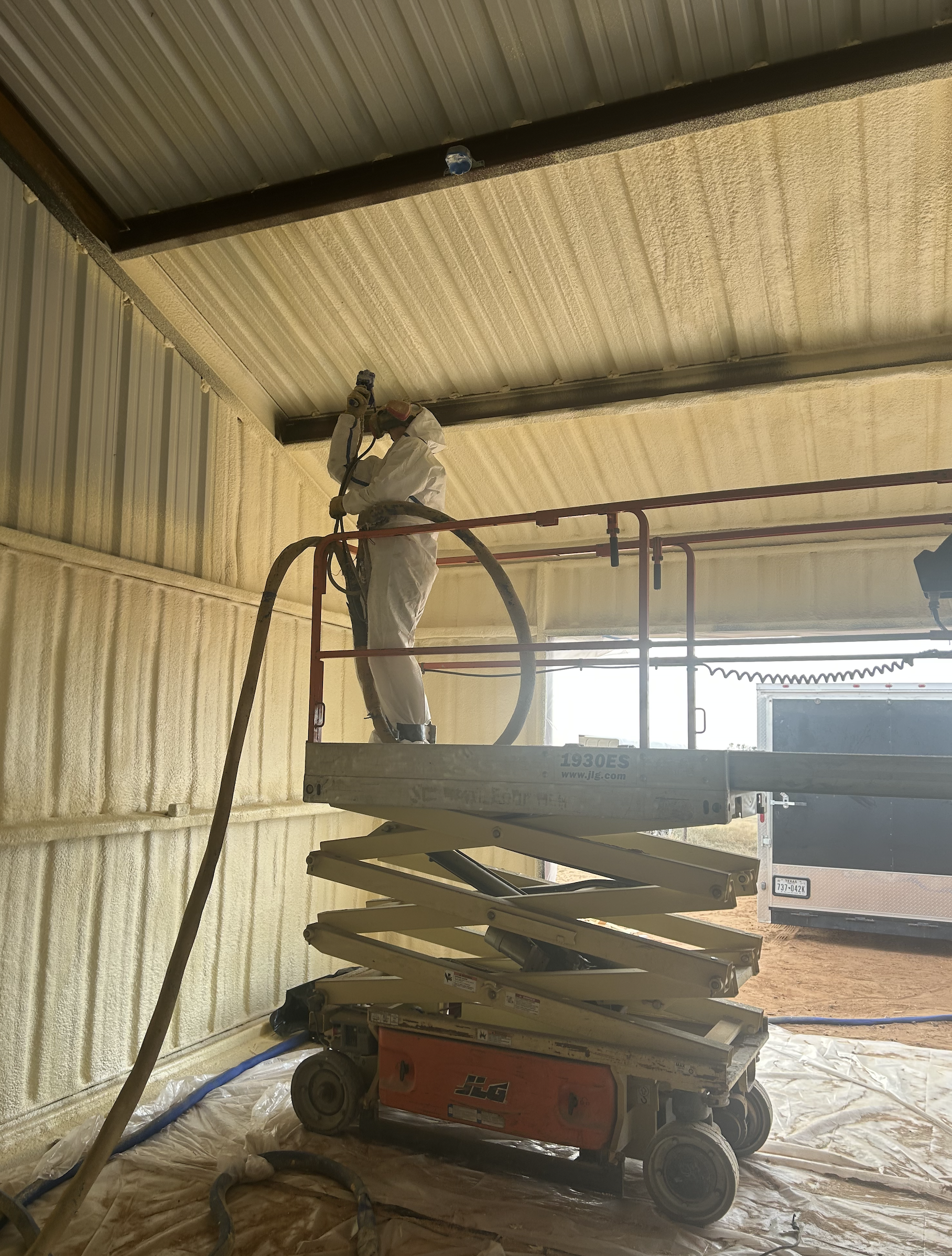 Worker in white protective suit and helmet spray-painting the metal ceiling of a building inside a scissor lift amid construction tools and materials.