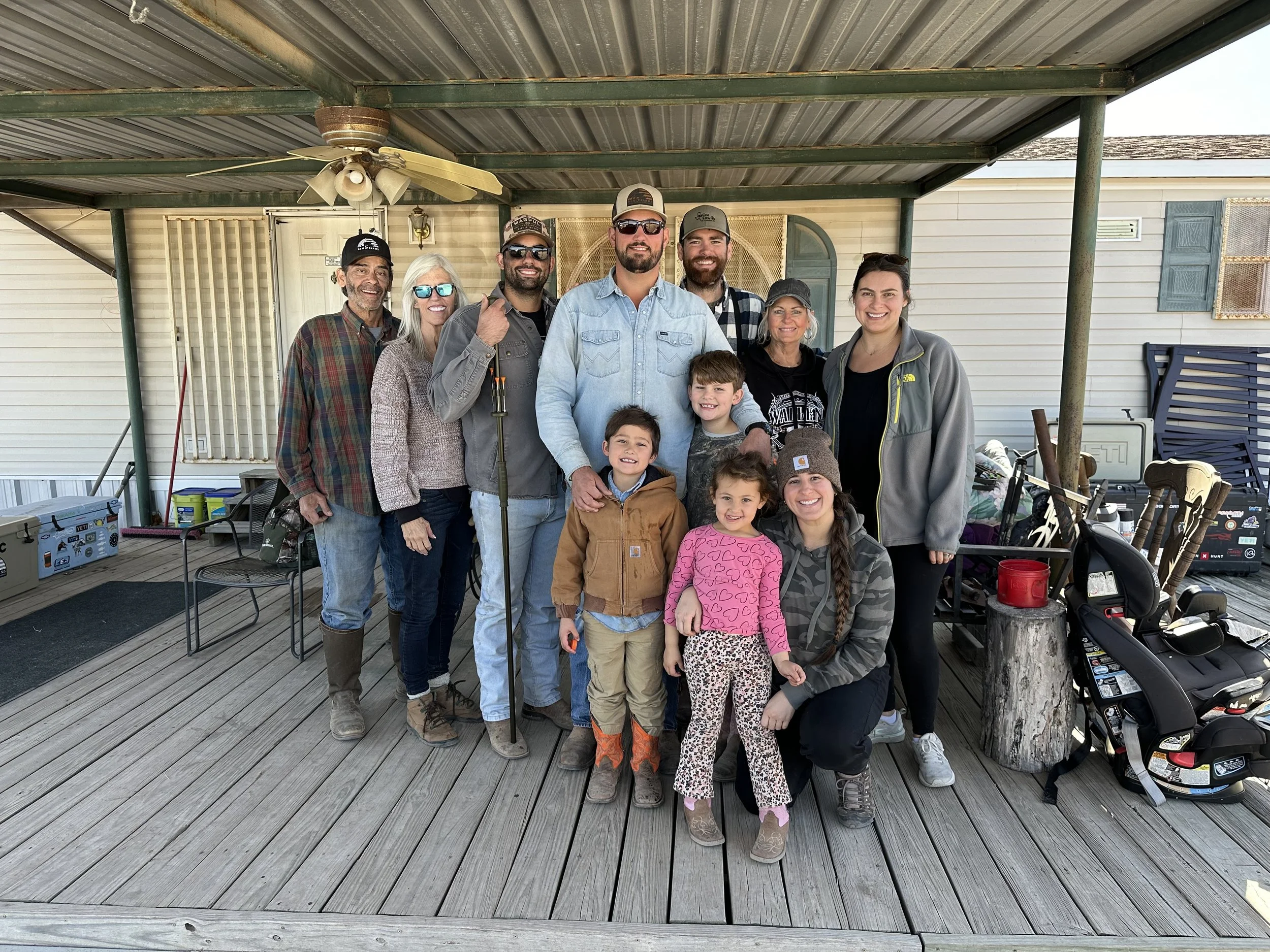 Group of people, including adults and children, smiling and posing for a photo on a wooden deck outside a house with a covered porch.