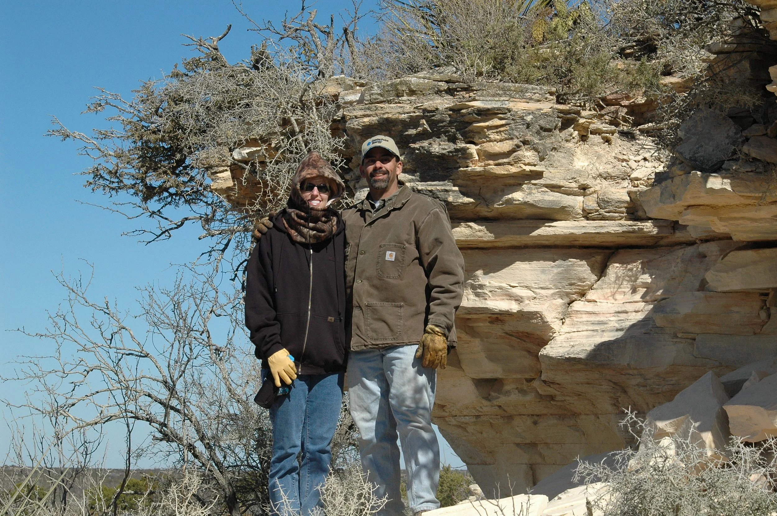 A man and woman are standing outdoors near large layered rock formations with leafless trees around them under a clear blue sky. The woman is wearing sunglasses, a hooded jacket, and gloves, and the man is wearing a cap, jacket, and gloves while smil