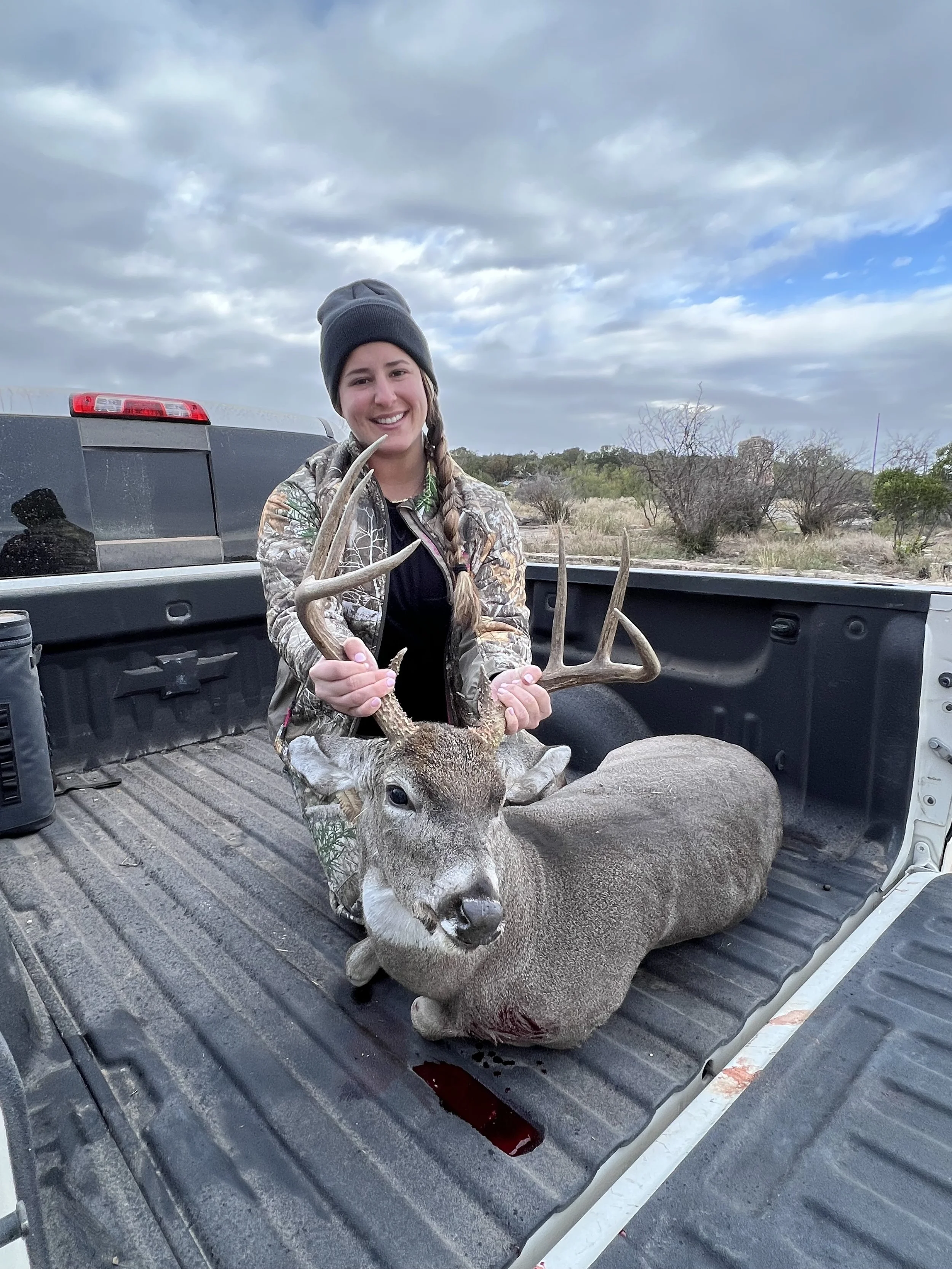 A woman in a camouflage jacket and beanie holding antlers, sitting on a large deer with a bloodstain on its neck, in the bed of a pickup truck in an open outdoor area with sparse trees and cloudy sky.