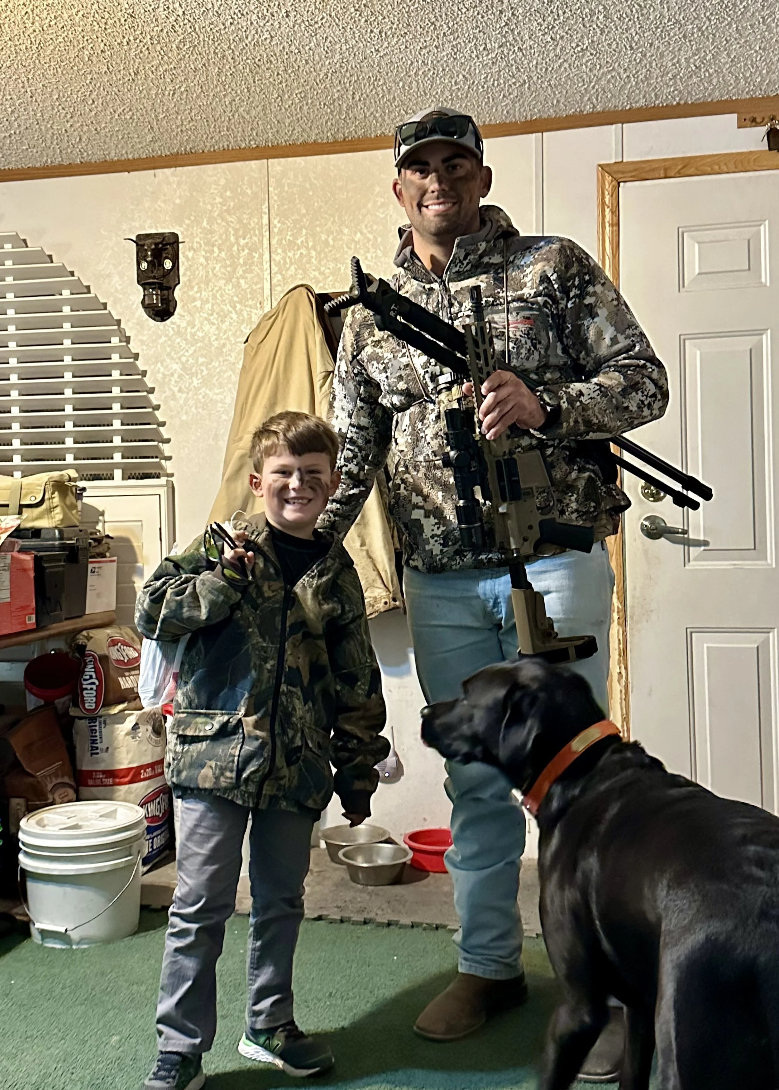 Two men, one young boy, and a black dog inside a room. The man on the right is holding a large firearm. The room has a green carpet, buckets, and bags of dog food.