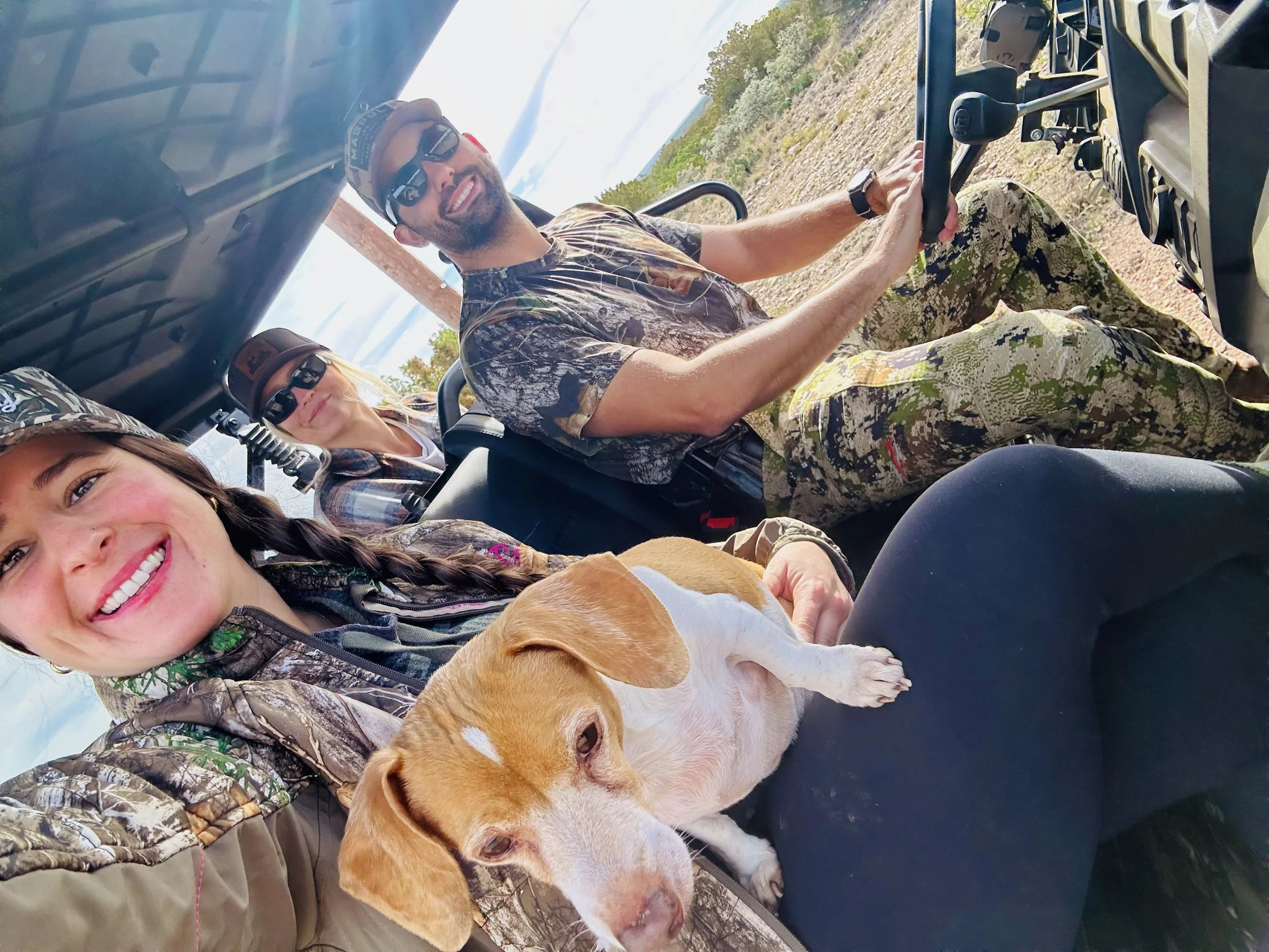 Group of people riding in an open vehicle, smiling, wearing camouflage clothing, with a dog in the foreground, outdoors with trees and a cloudy sky.