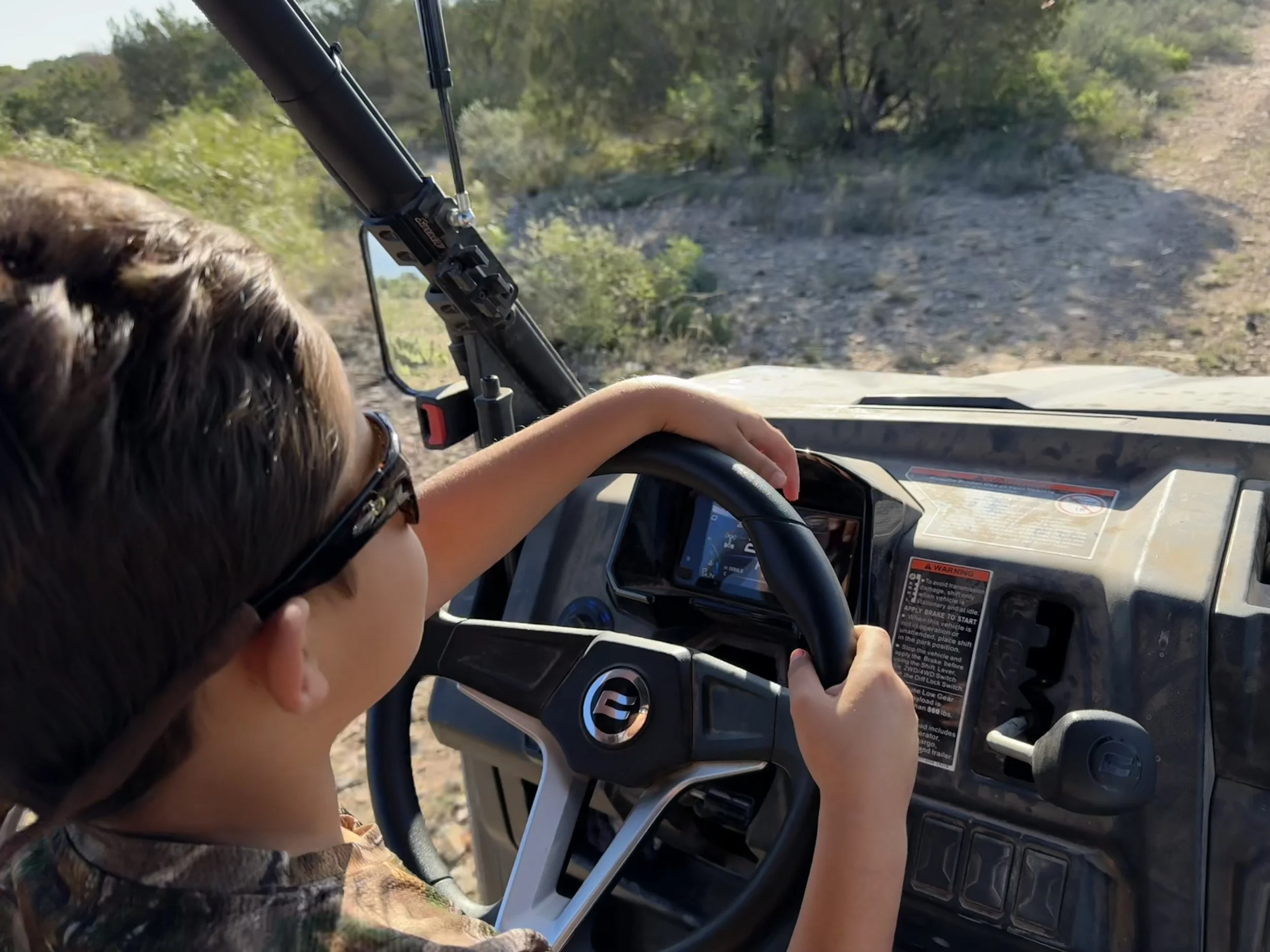 Child driving a utility vehicle on a dirt trail surrounded by greenery, with the child's arm resting on the steering wheel and wearing sunglasses.