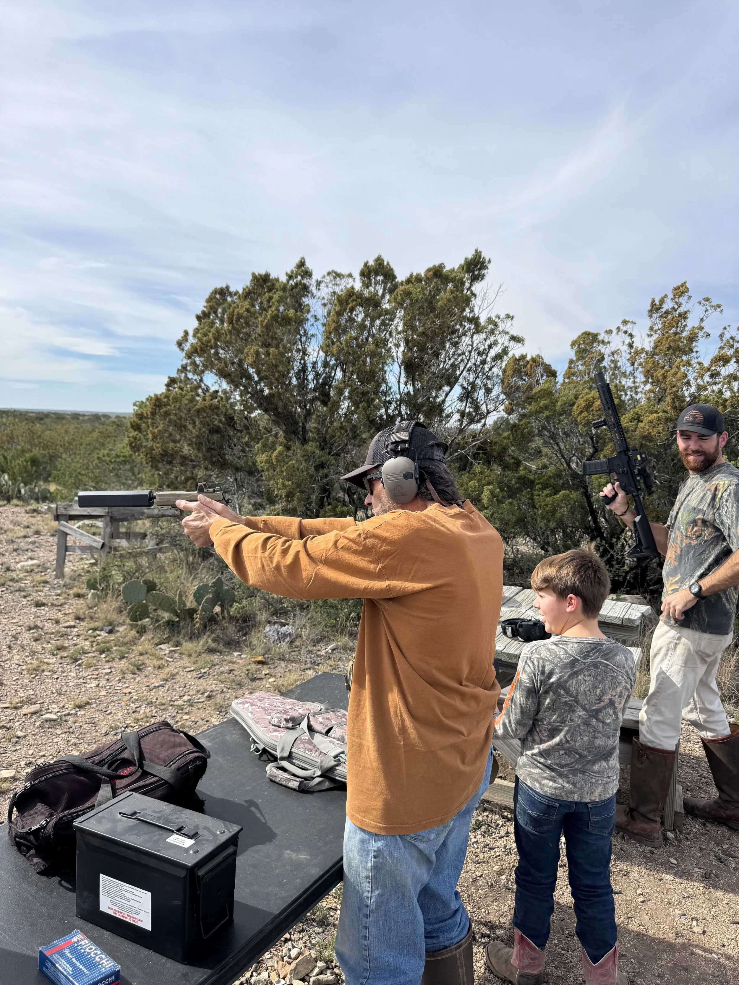 Man aiming a firearm at an outdoor shooting range, with two children observing, trees and a cloudy sky in the background, and shooting gear and equipment on a table.