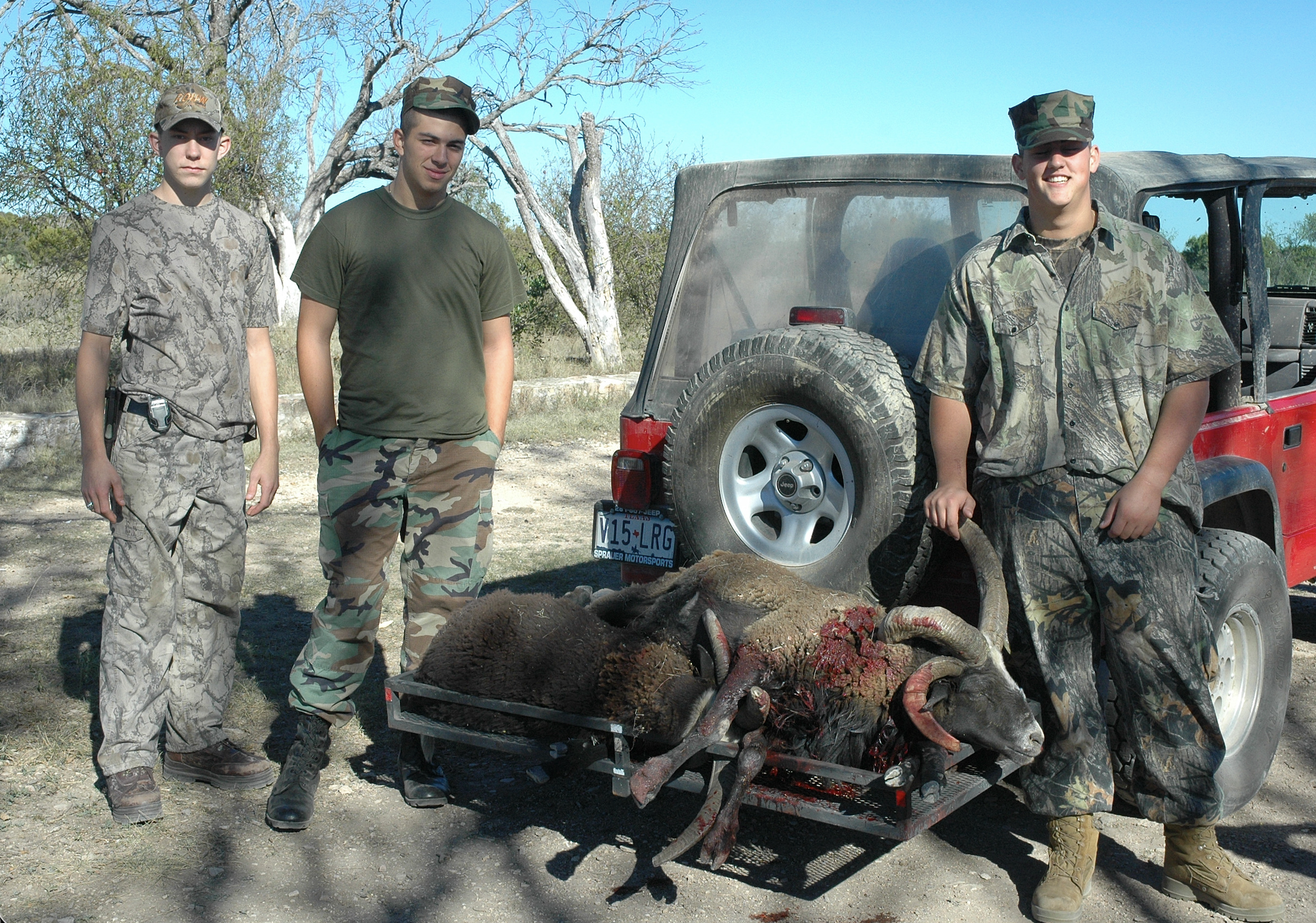 Three young men in camouflage and military-style clothing stand outdoors next to a red off-road vehicle, with a carcass on a cart in front of them, in a dry, wooded area.